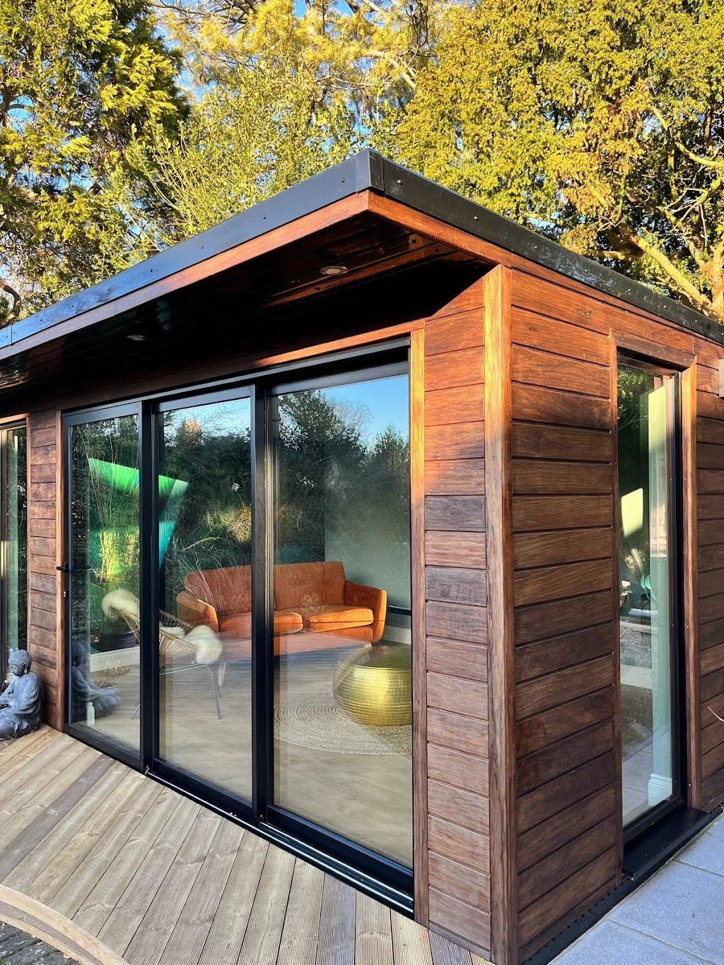 A modern wooden house with large glass sliding doors. Inside, there is an orange couch and a round golden coffee table. The house is surrounded by trees and is built on a wooden deck.