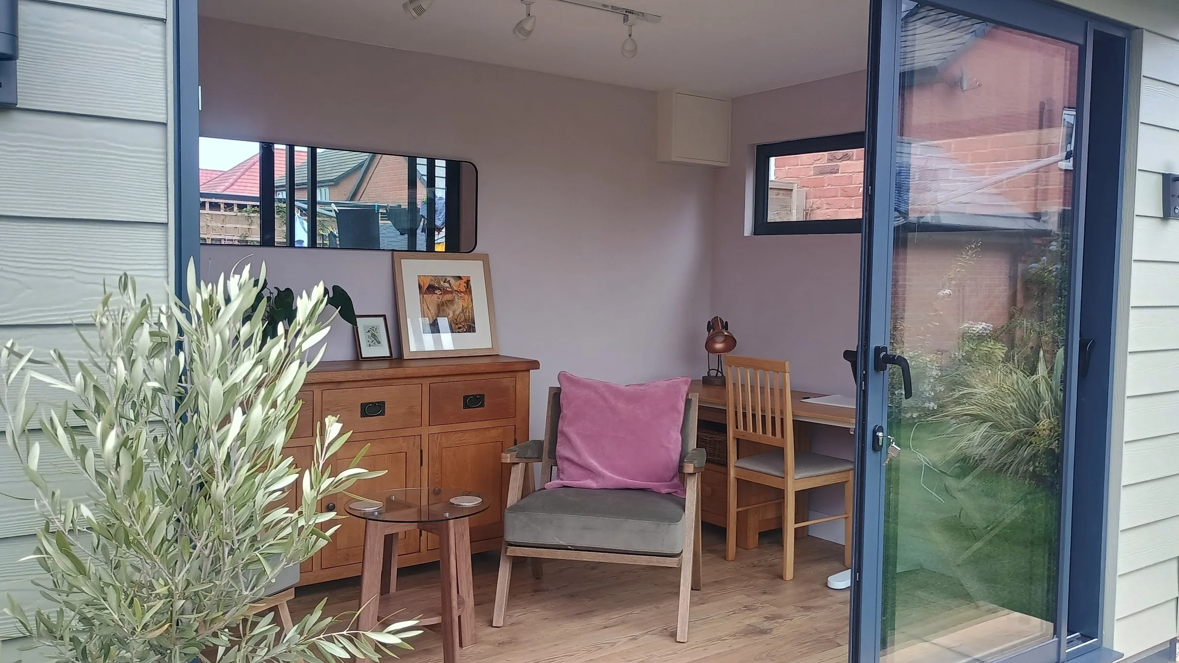 Interior view of a cozy room with wooden furniture, including a sideboard with framed artwork, a gray armchair with a pink cushion, a small round glass-top table, a desk with a wooden chair and desk lamp, and large windows providing natural light and