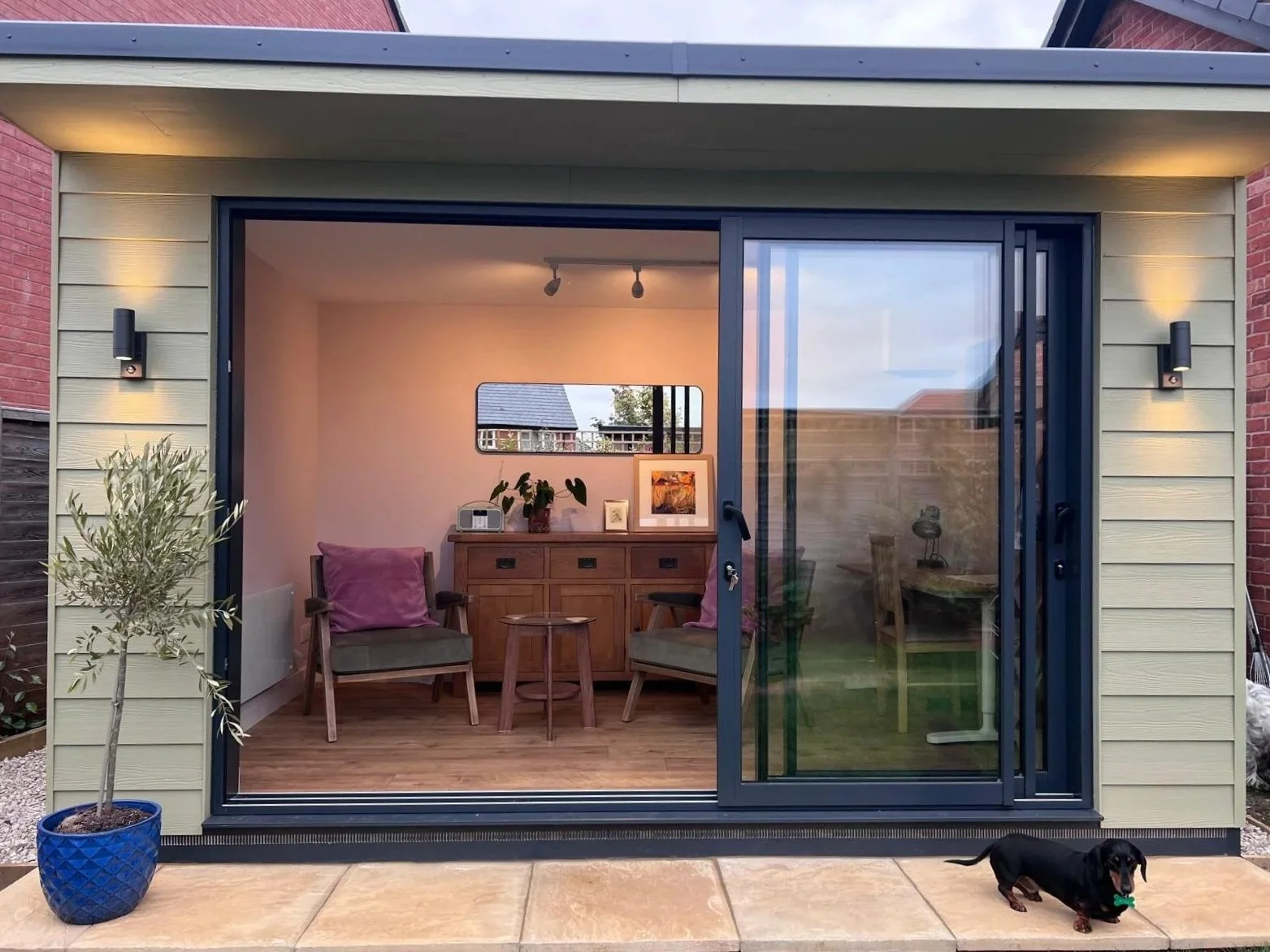 View of a modern small backyard extension with large sliding glass doors revealing a cozy indoor seating area with two armchairs, a small table, a sideboard, and decorative items. A potted plant is outside on the patio, and a small black Dachshund pu