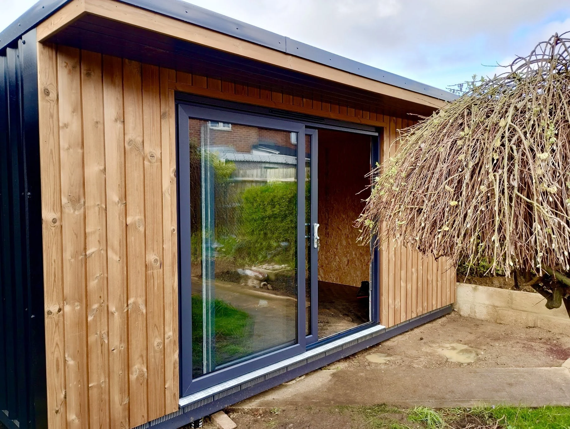 A small modern garden shed with wooden and black metal panels, a large glass sliding door, and a nearby tree with drooping branches