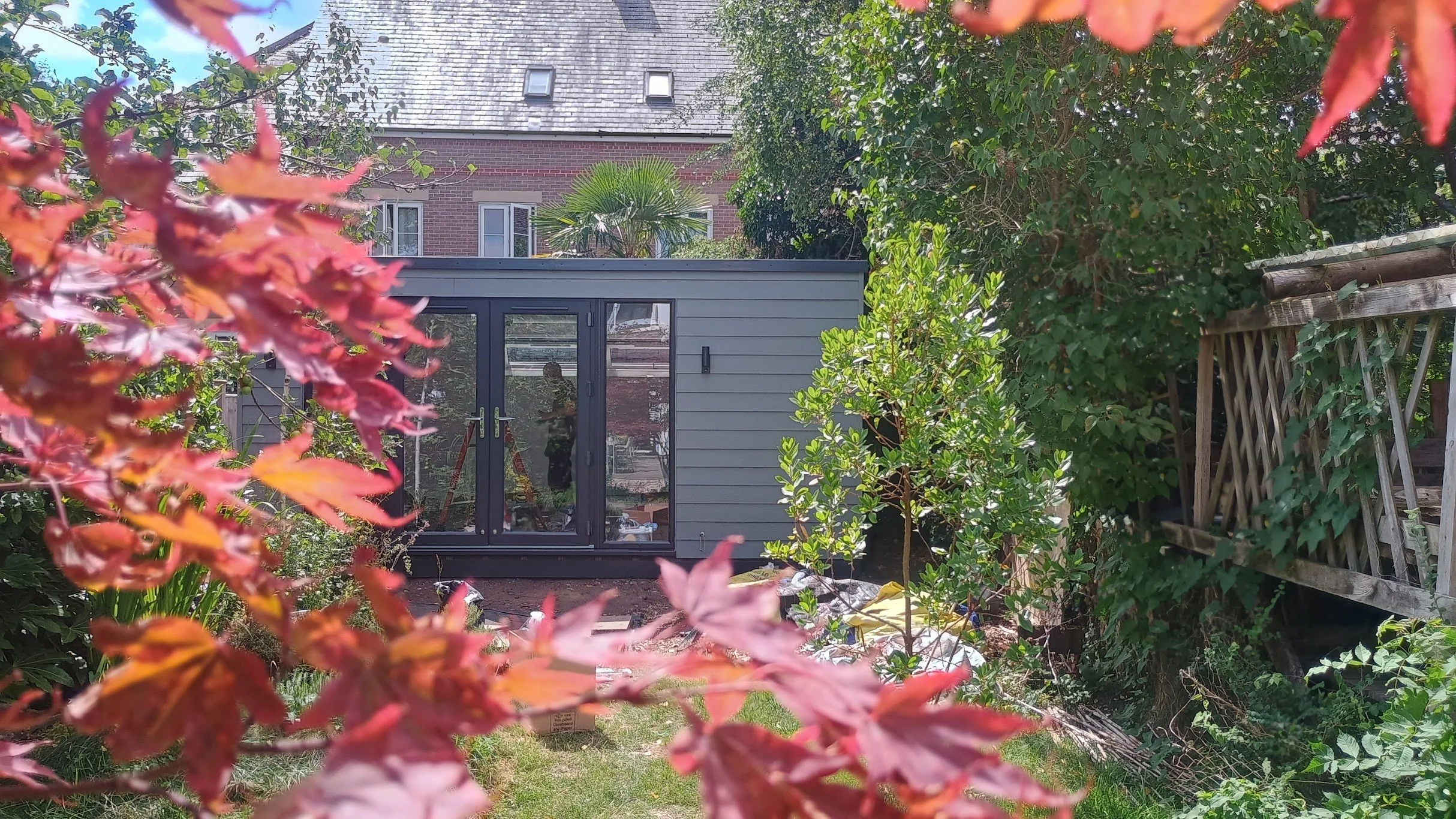 A backyard garden viewed through vibrant red and green leaves, with a grey garden room featuring glass double doors and a small outdoor area surrounded by lush greenery, a wooden fence on the right, and a multi-story brick house with a steep shingled