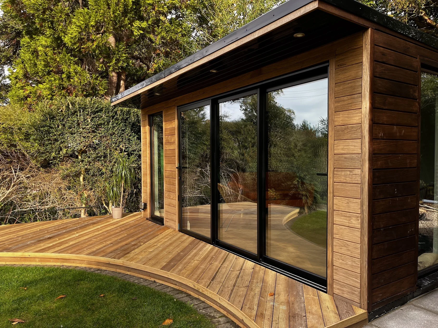 Small modern wooden house with large glass sliding doors, surrounded by trees and a wooden deck with a small section of grass in the foreground.