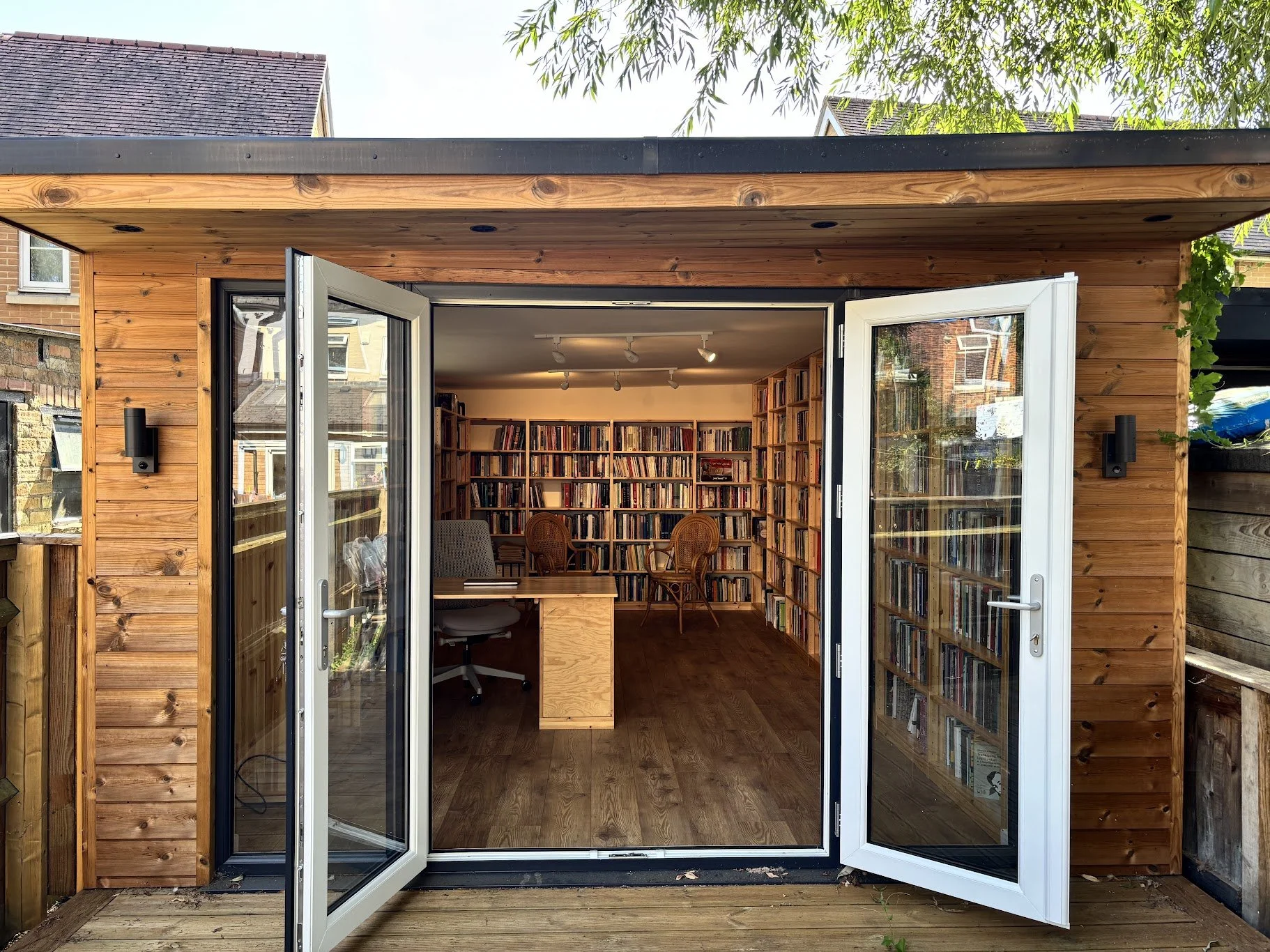 View of a small wooden shed with open glass double doors, revealing a cozy interior with bookshelves filled with books, a desk with a swivel chair, and several wooden chairs, surrounded by a backyard with leafy trees.