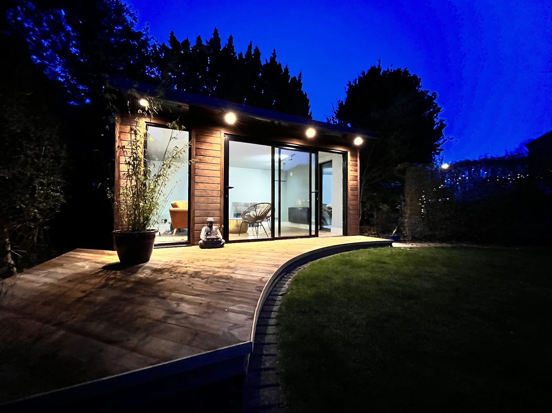 A small wooden house with large glass doors, illuminated by outdoor lights, sits on a wooden deck surrounded by trees and a grassy yard at night.
