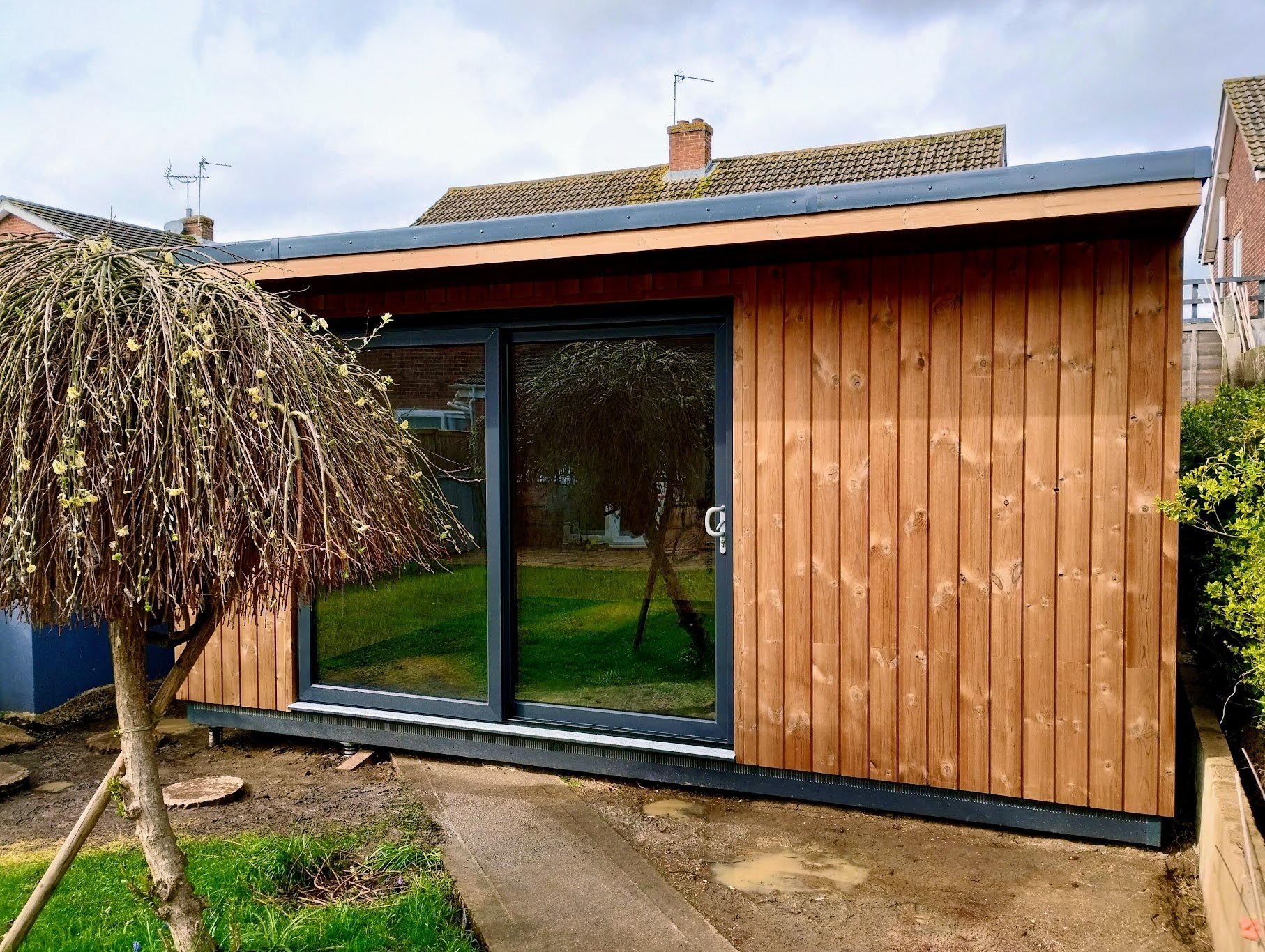 Small wooden garden shed with large sliding glass door, partially paved backyard with a small tree and green grass, neighboring houses visible in the background, and an overcast sky overhead.