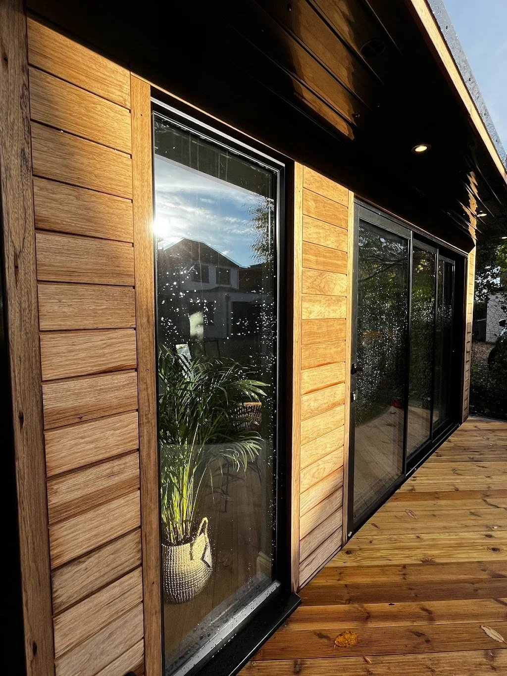 A wooden house exterior with large glass sliding doors, a wooden deck, and a potted plant inside reflected in the glass, with sunlight and clouds visible through the glass.