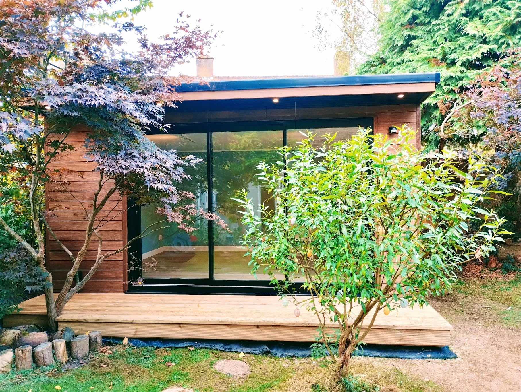 A small modern house with a wooden exterior, large glass sliding doors, and a wooden deck. There are trees and bushes surrounding the house with purple and green leaves.