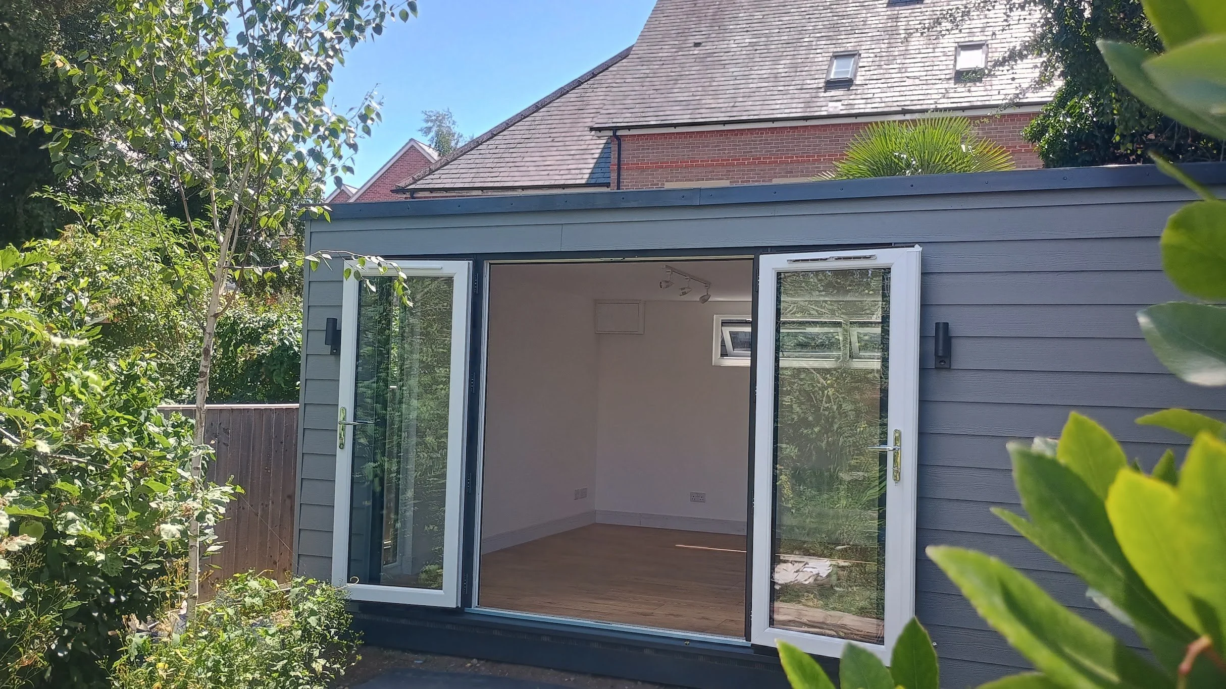 Small gray garden room with open double glass doors, interior with white walls, small window, and wooden floor, surrounded by green foliage, under a blue sky.