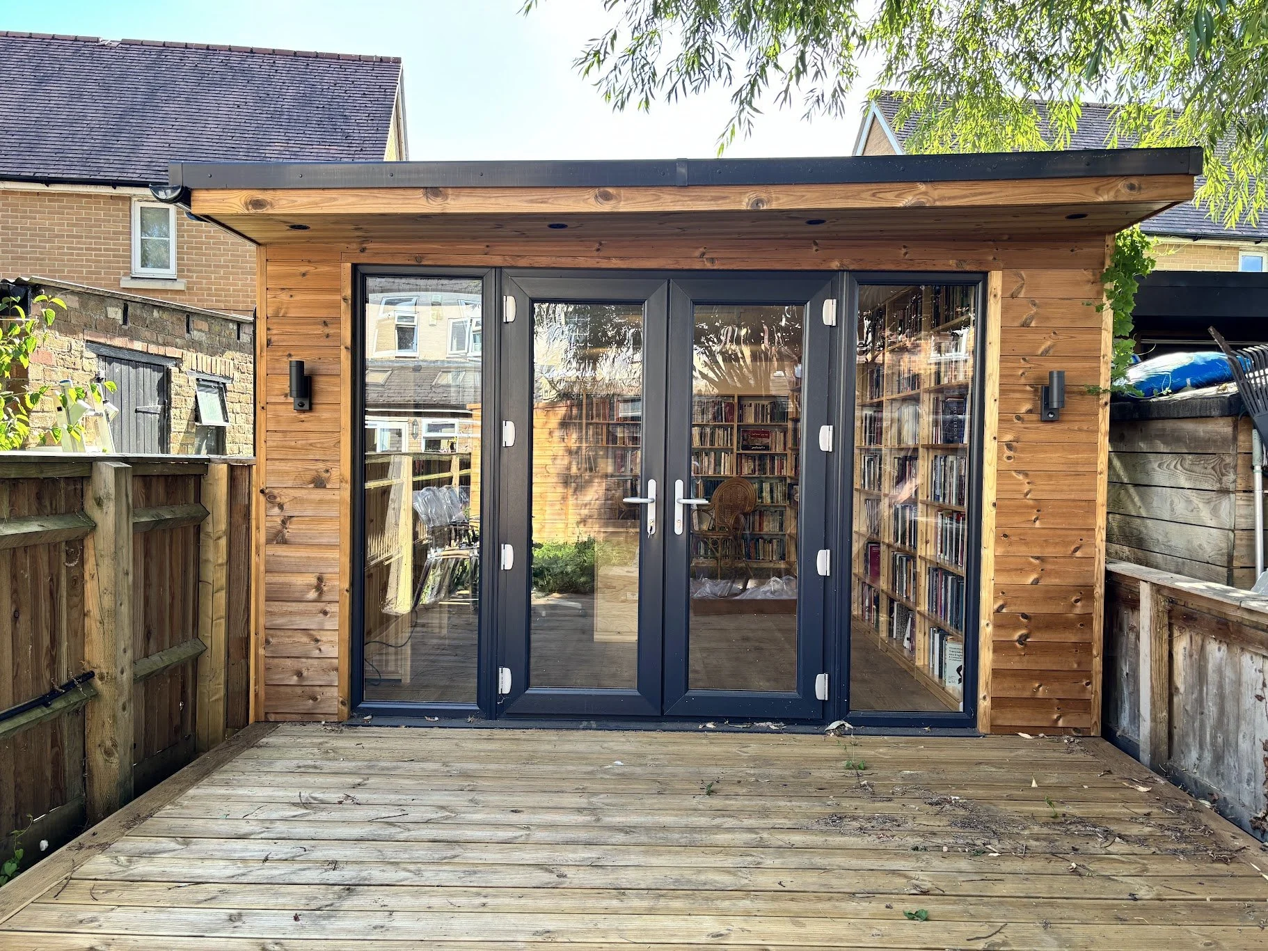 Backyard wooden shed with large glass double doors and bookshelves visible inside, located on a wooden deck with neighboring brick houses and greenery.