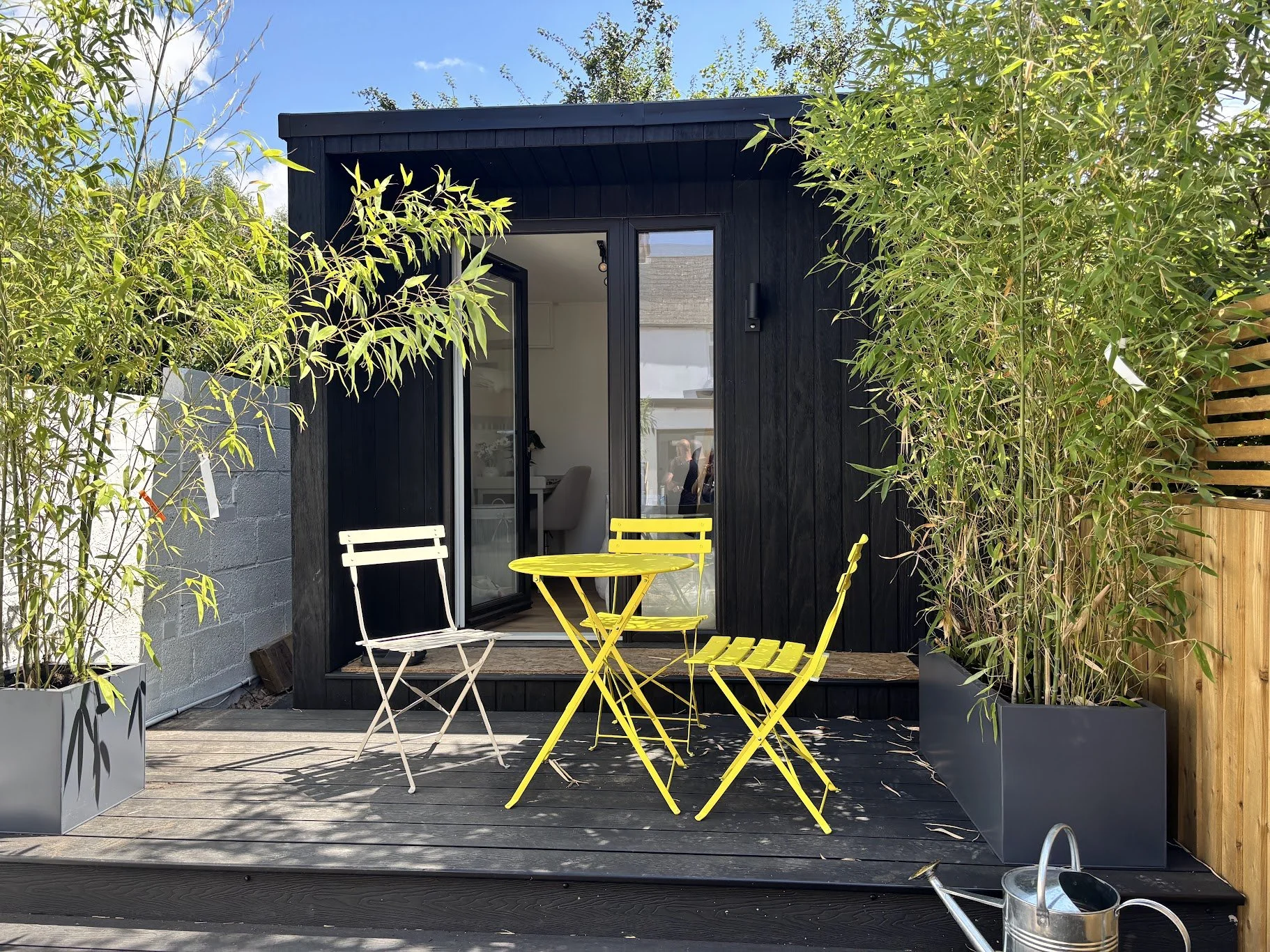 A small outdoor patio with yellow and white metal chairs and a matching yellow table in front of a black wooden tiny house, surrounded by potted bamboo plants and a wooden fence.