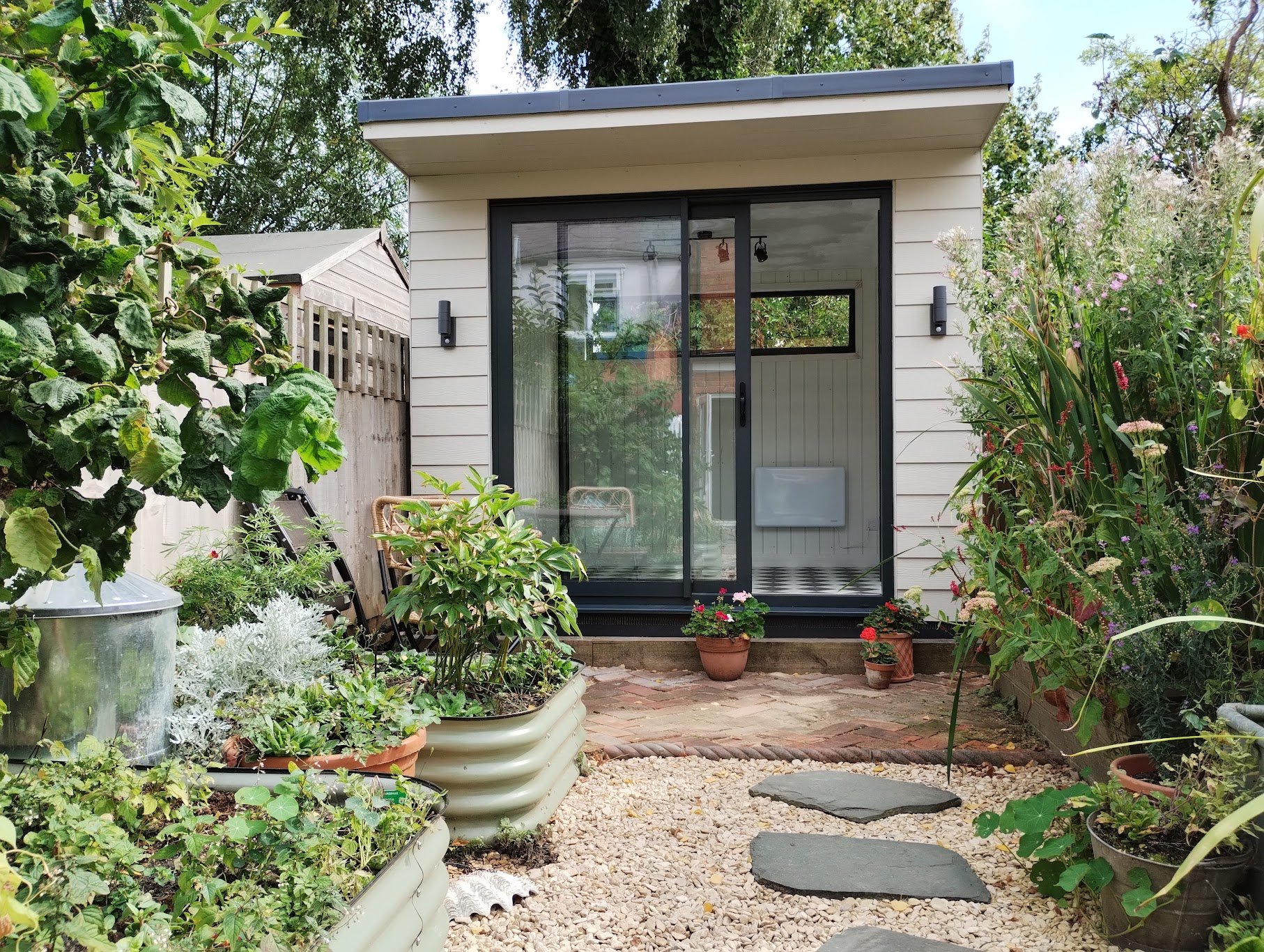 A small backyard garden with potted plants, a gravel path with stepping stones leading to a glass sliding door, surrounded by lush greenery and flowering plants.