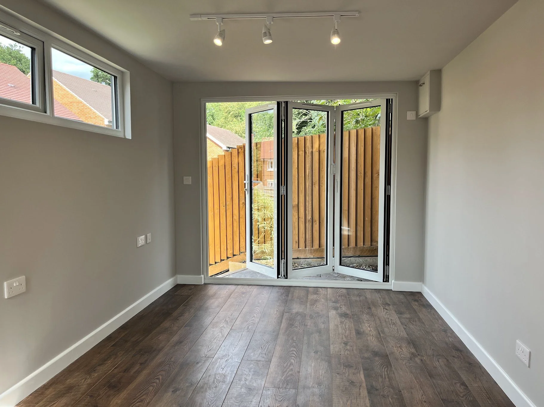 Empty room with wooden floor, gray walls, small horizontal window, and glass door leading outside with wooden fence.