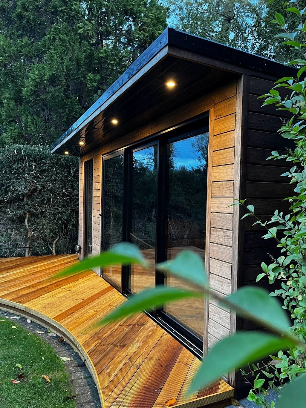 A modern tiny house with large glass sliding doors, wooden exterior, and small overhang with recessed lighting, surrounded by greenery at dusk.