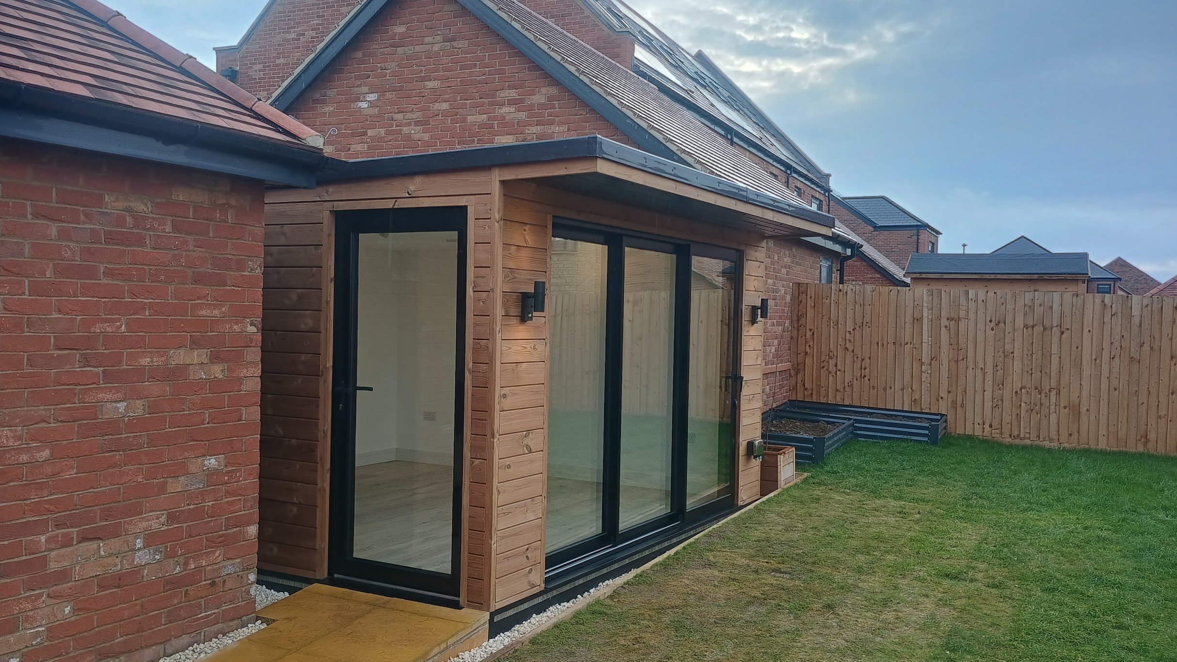 Backyard with a brick house and a wooden extension with large glass sliding doors, a lawn, and a wooden fence.