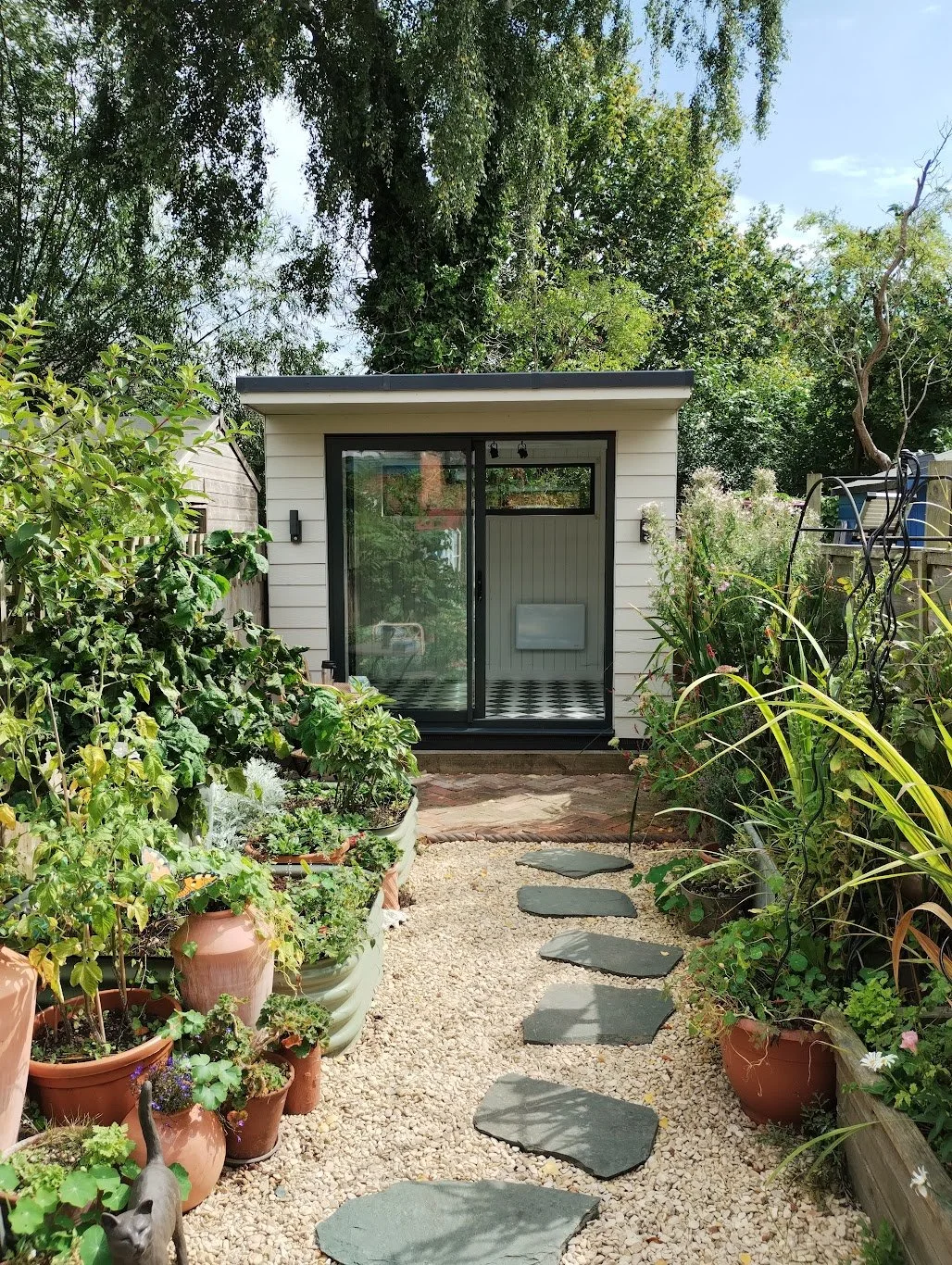 A small garden with potted plants on both sides and a gravel pathway with stepping stones leading to a small outbuilding with large glass doors and a window, surrounded by greenery.