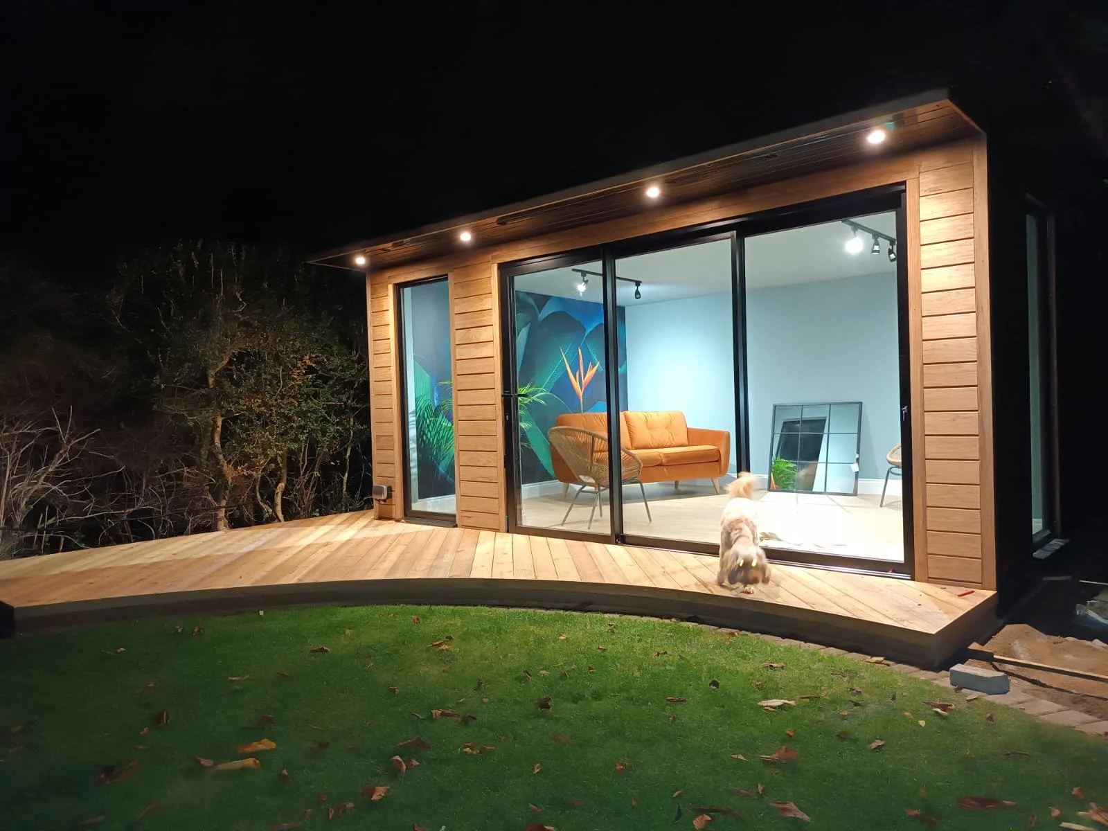 A small modern house with large glass sliding doors, a wooden deck, and a green lawn at night. Inside, there is furniture, a large mirror, and a mural on the wall. A dog is sitting outside on the deck, looking into the house.
