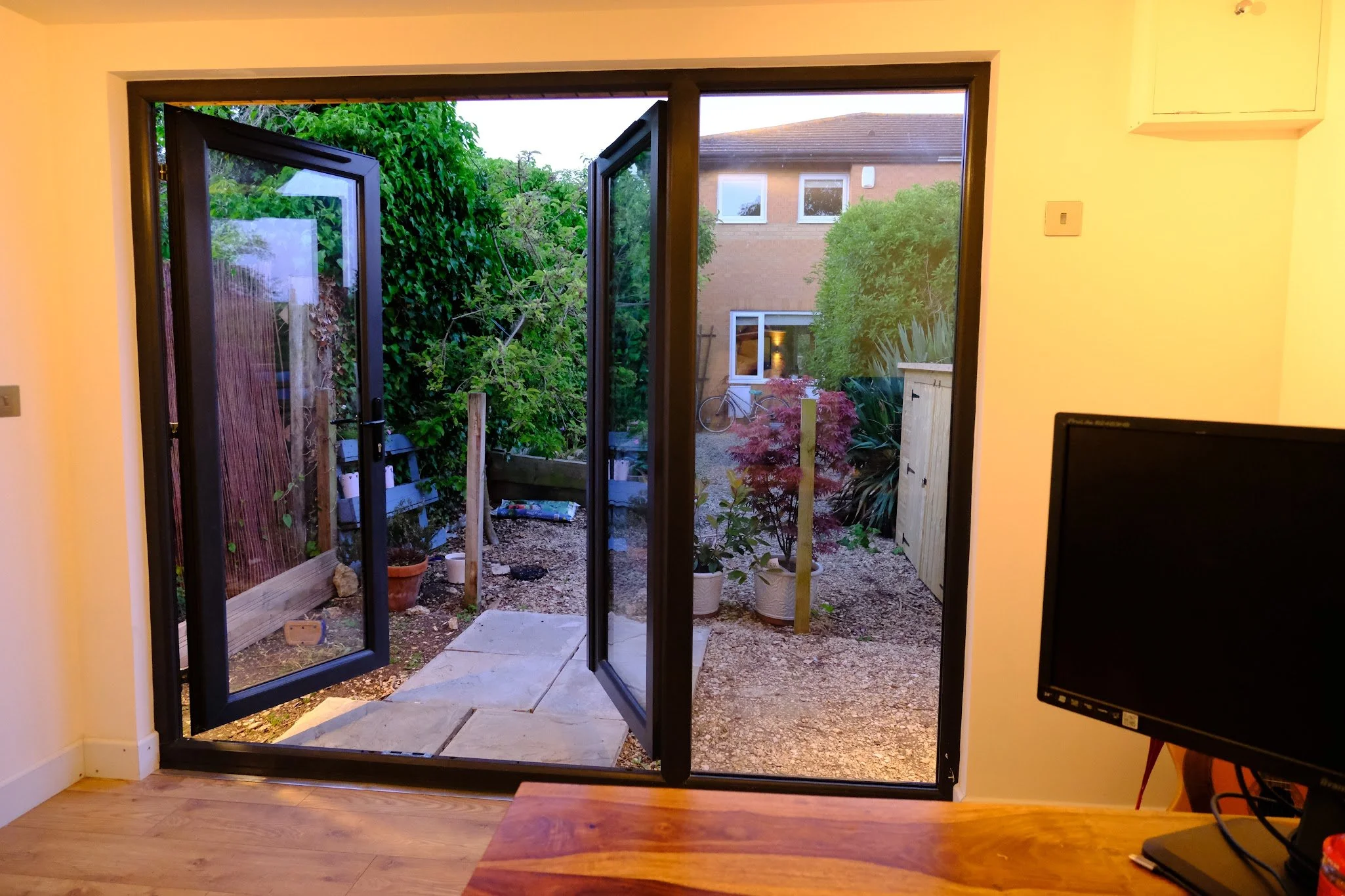 Looking through open glass double doors into a backyard garden with plants, bushes, a bicycle, and neighboring houses.