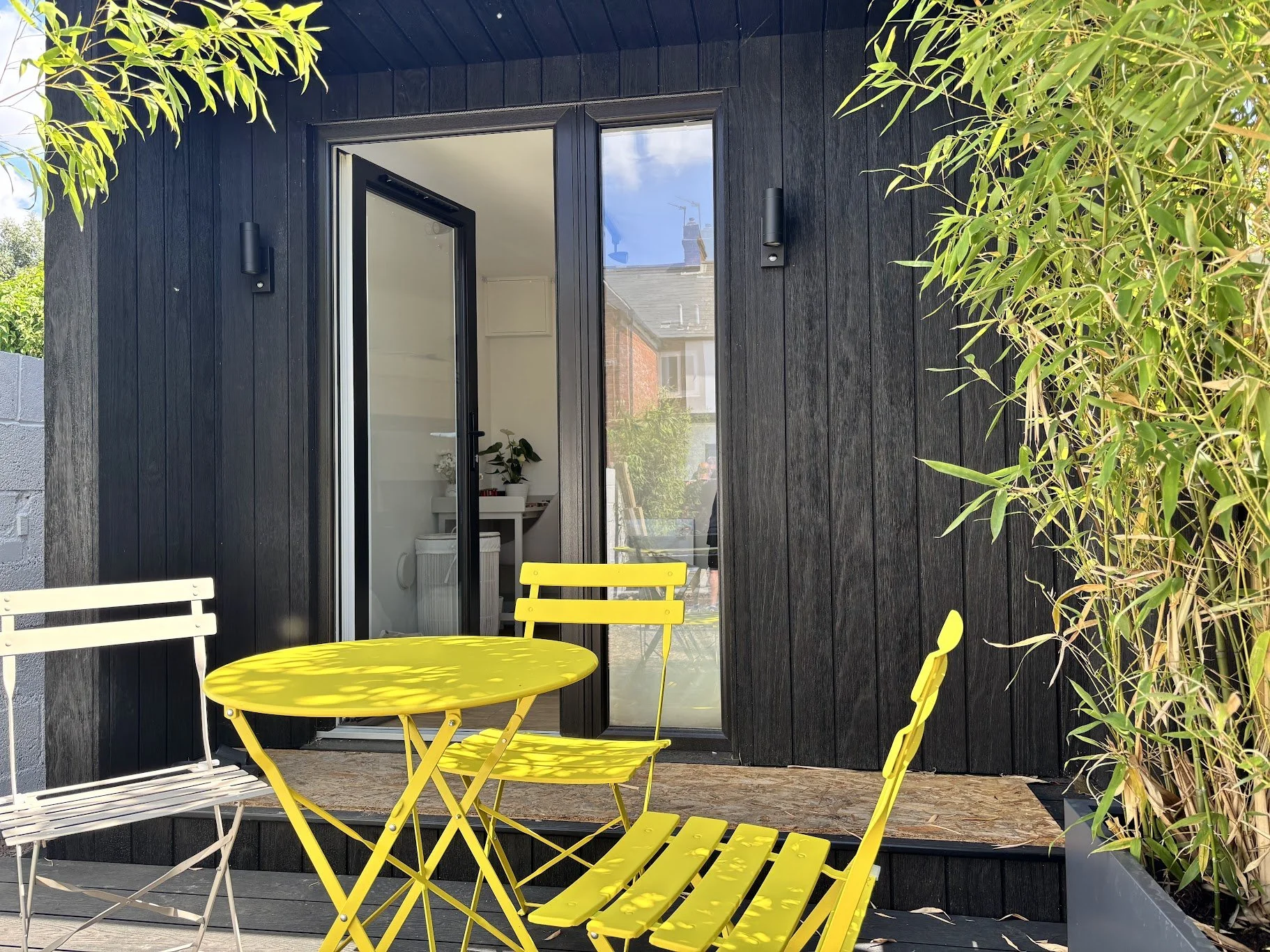 Black wooden exterior wall of a house with a glass door, leading to an interior room with a white desk and plants. Outdoor patio area with yellow and white metal furniture, including a round table and chairs, and potted bamboo plants on the right.