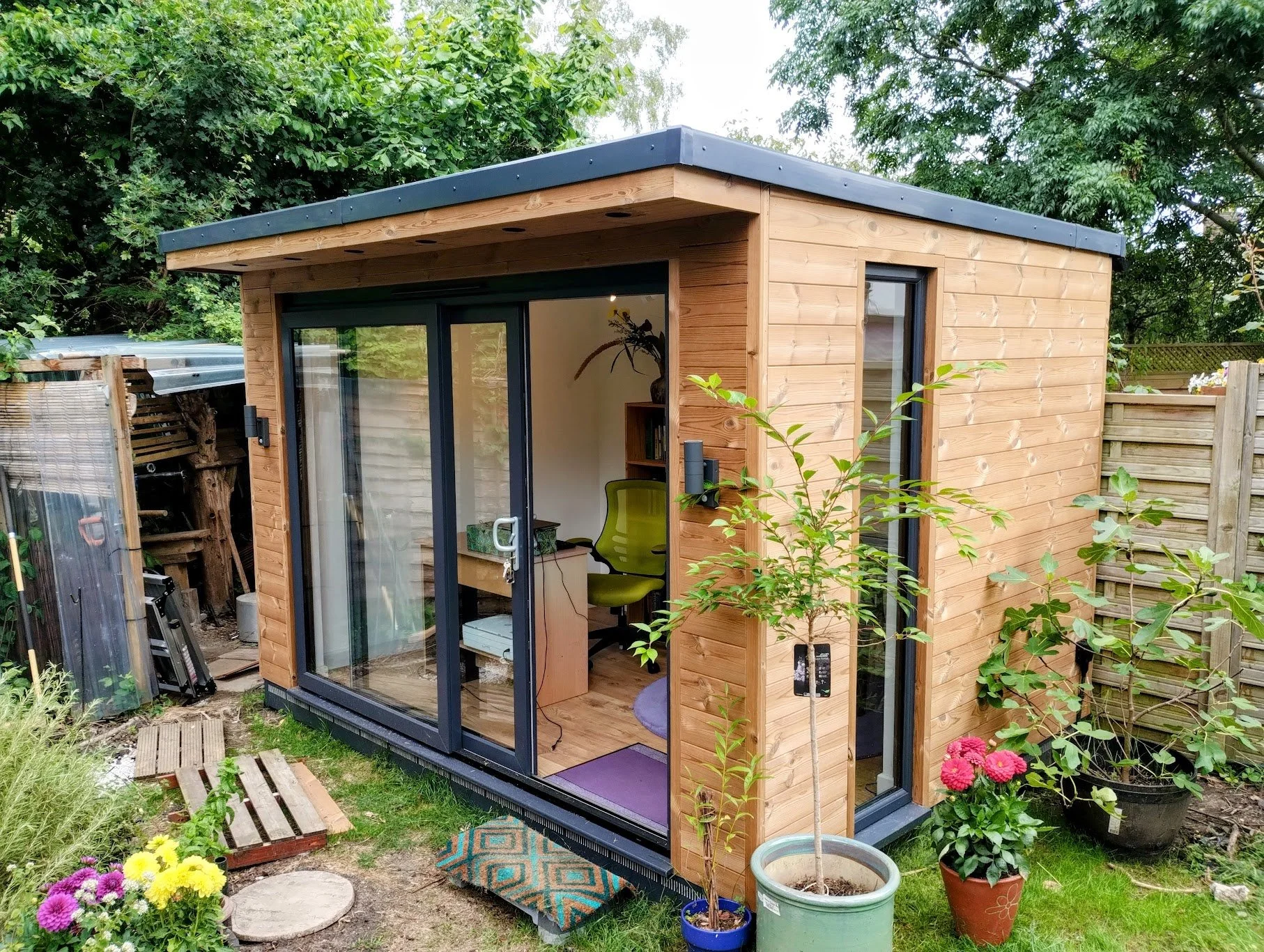 Small wooden garden office with glass sliding doors and narrow side window, surrounded by potted plants and flowering bushes, with tools and a small path nearby.