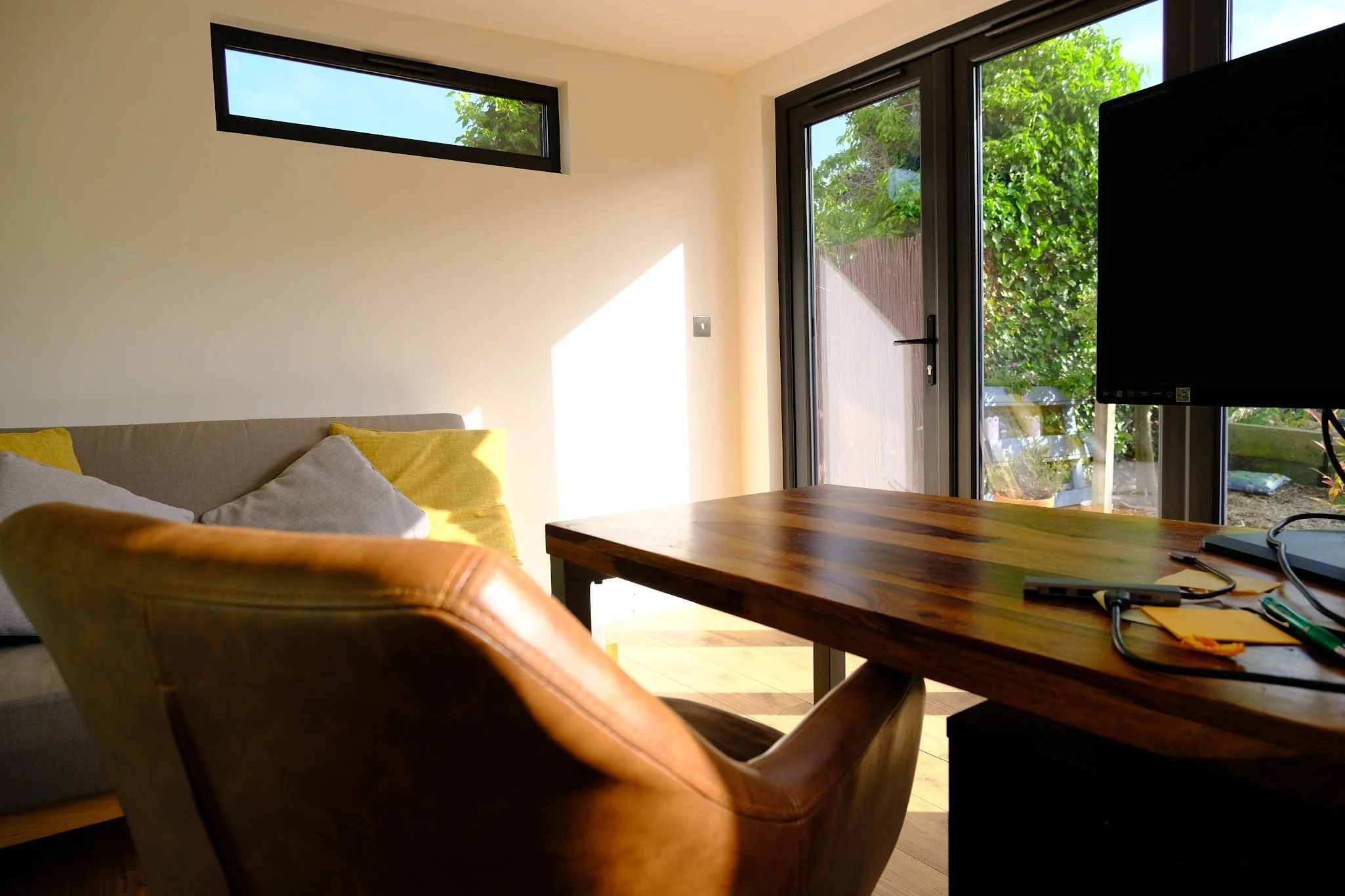 A cozy living room with a brown leather armchair, a beige couch with yellow and white pillows, a wooden table, a wall-mounted TV, and large windows letting in natural sunlight, revealing greenery outside.