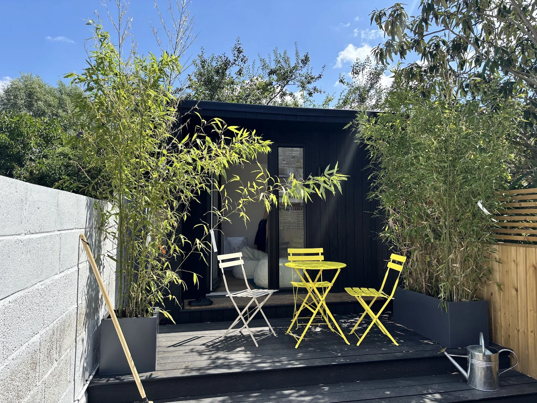 Small outdoor patio with a black shed, yellow and white chairs and table, bamboo plants in pots, and a watering can under a blue sky with clouds.