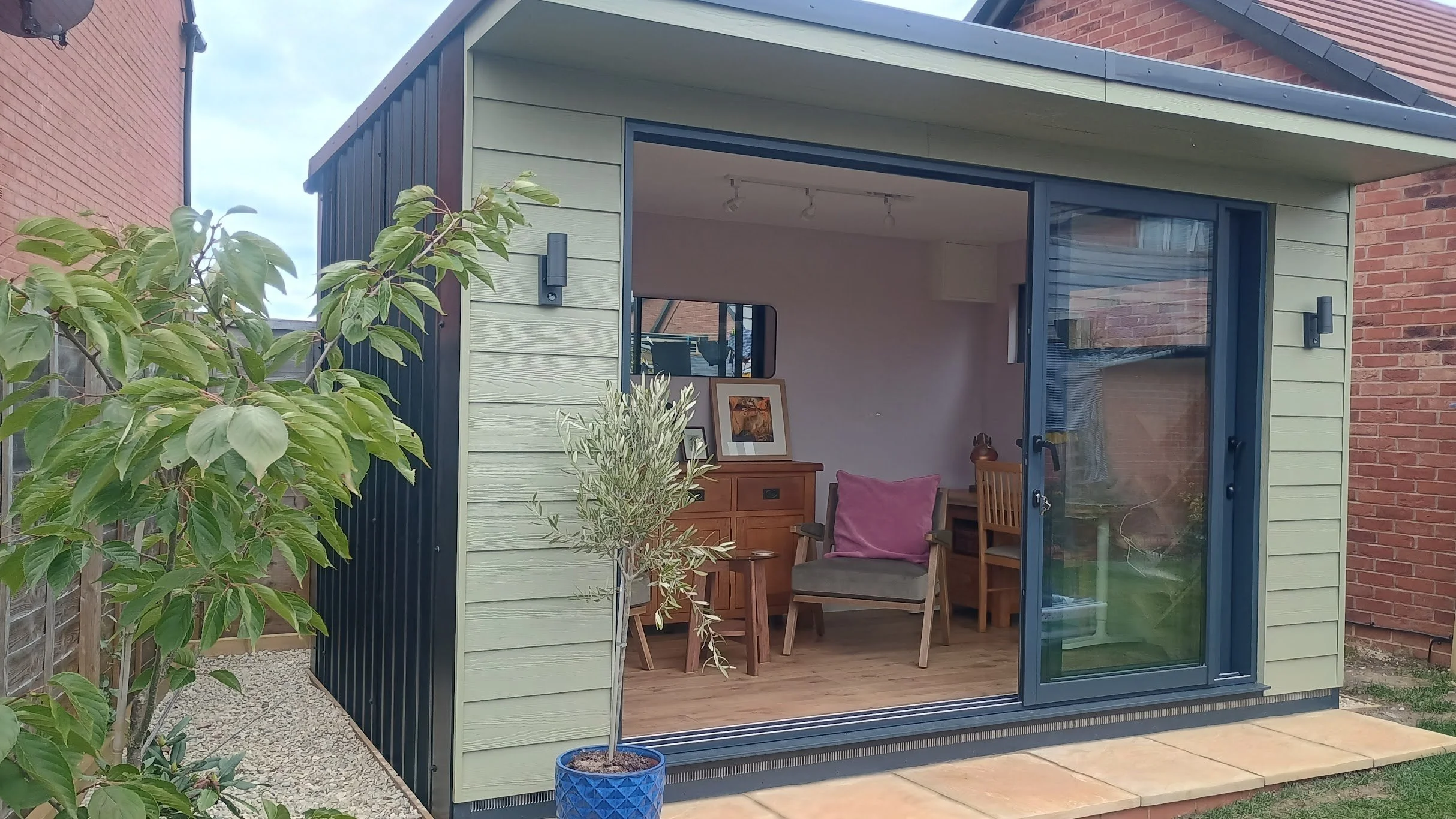 Small backyard shed with open sliding glass door, showing a cozy interior with a chair, cushion, and framed photos on a wooden sideboard. A potted plant is outside the shed, with a gravel pathway and brick wall nearby.