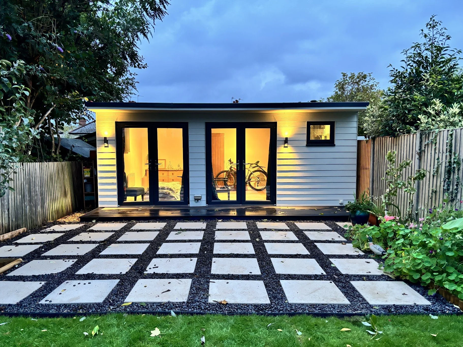 Backyard with small modern white shed, black-framed glass doors, potted plants, garden bed, and patterned stone patio at dusk.