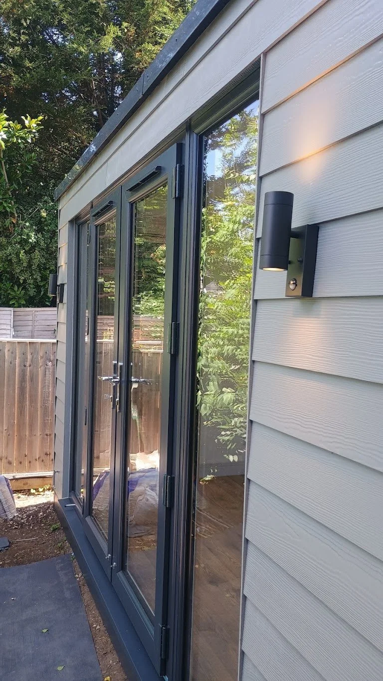 Exterior view of a house with sliding glass doors, beige siding, and a black wall-mounted outdoor light fixture, with trees and a wooden fence in the background.