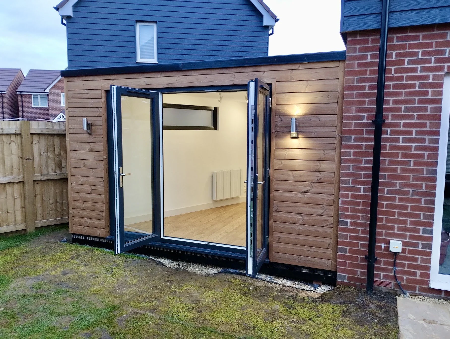 Small backyard wooden garden room with glass double doors, mounted on a metal base, between a red brick wall and a wooden fence, with a grassy ground.