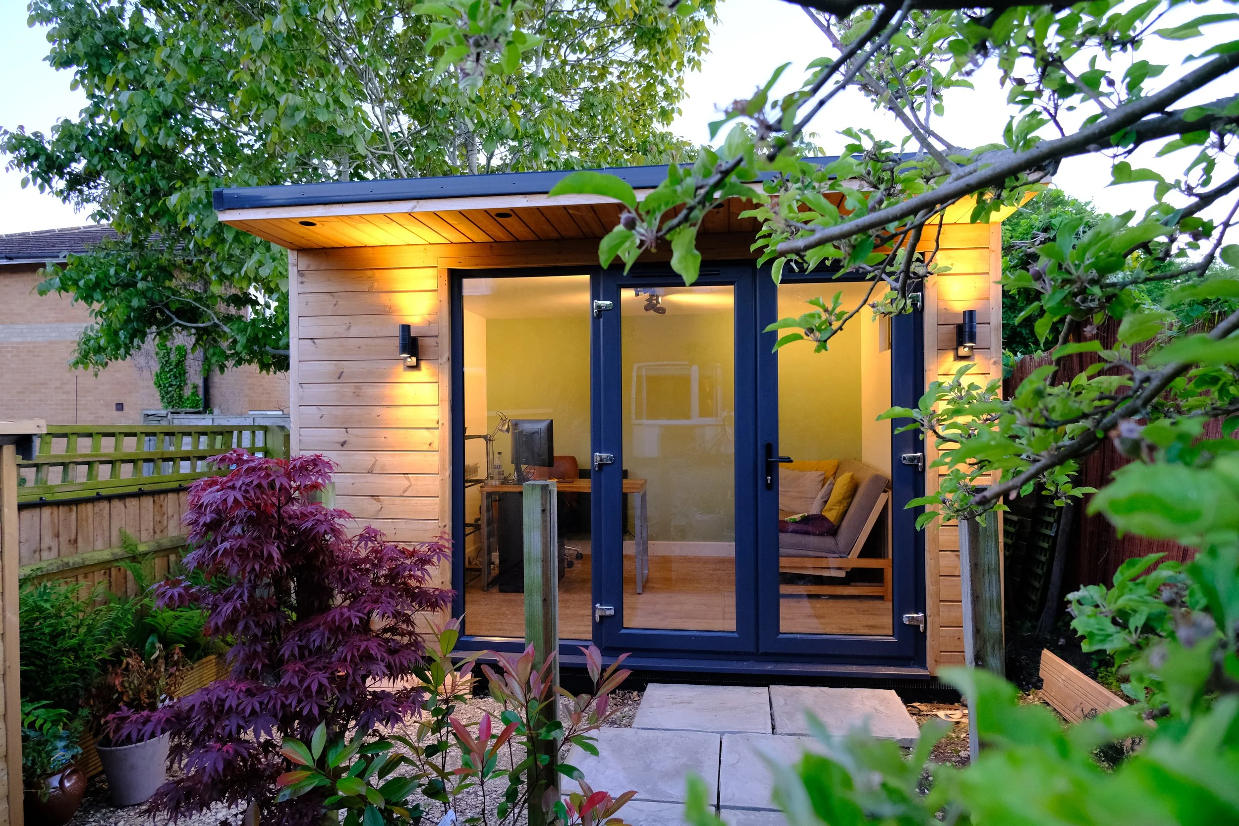 A small wooden garden shed with glass doors and warm interior lighting, surrounded by plants and trees.