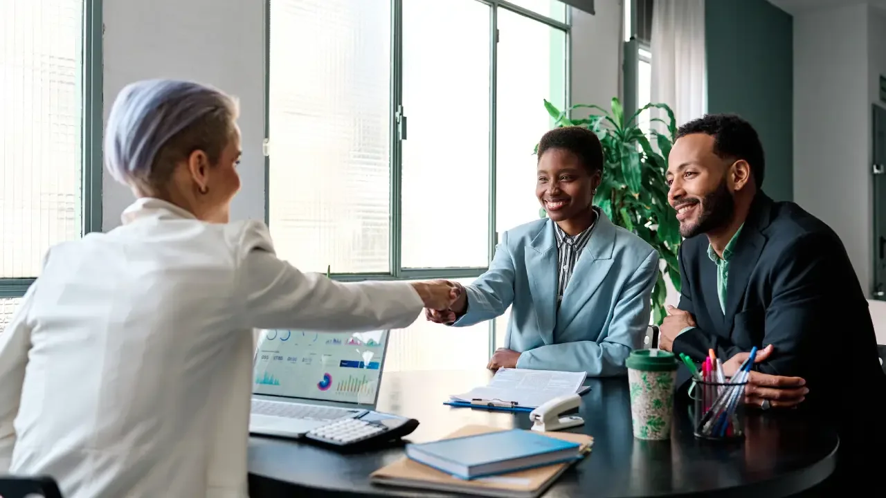 Two women and a man at a business meeting, shaking hands, smiling, in an office with large windows, notebooks, and coffee cups on the table.