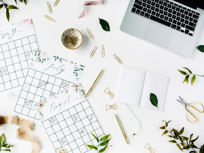 A white desk with a silver laptop, several open planners for July and August, gold pens, paper clips, scissors, green leaves, a small bowl filled with push pins, and other stationery items arranged neatly.