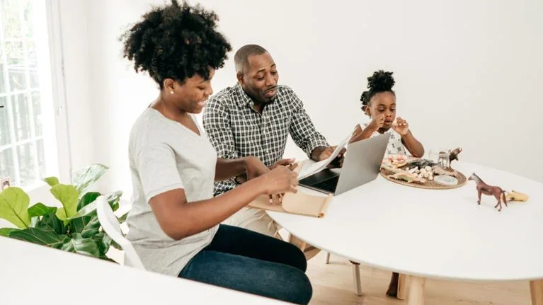 A family of three sitting at a white dining table, engaging with a laptop and looking at a book, with toys and a plant nearby.