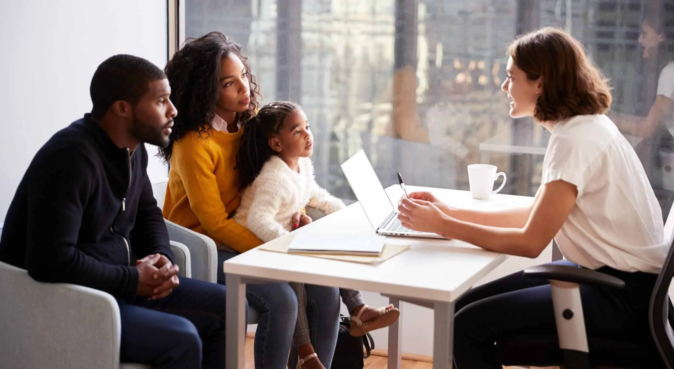 A woman consulting with a family of four, seated in a bright office with large windows, as she takes notes on a laptop.