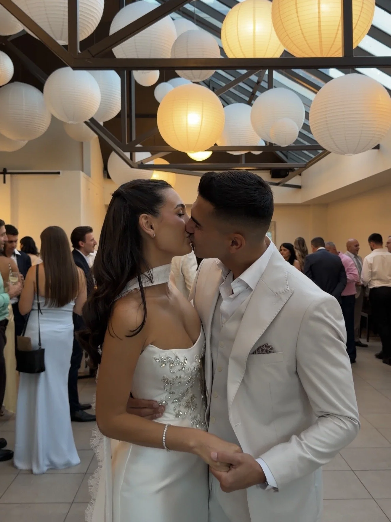 A bride and groom kissing at a wedding reception, with guests in the background and paper lanterns hanging from the ceiling.