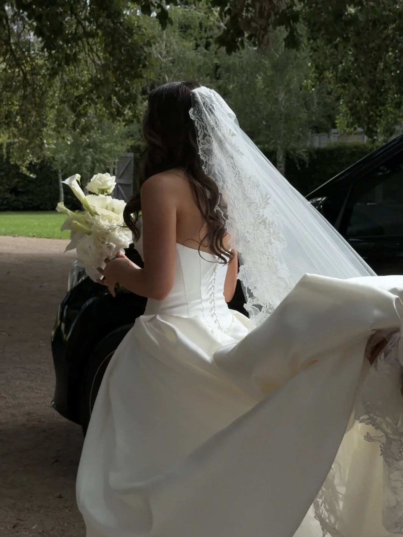 Bride in a white wedding dress with lace veil holding a bouquet of white flowers, standing beside a black car outdoors.