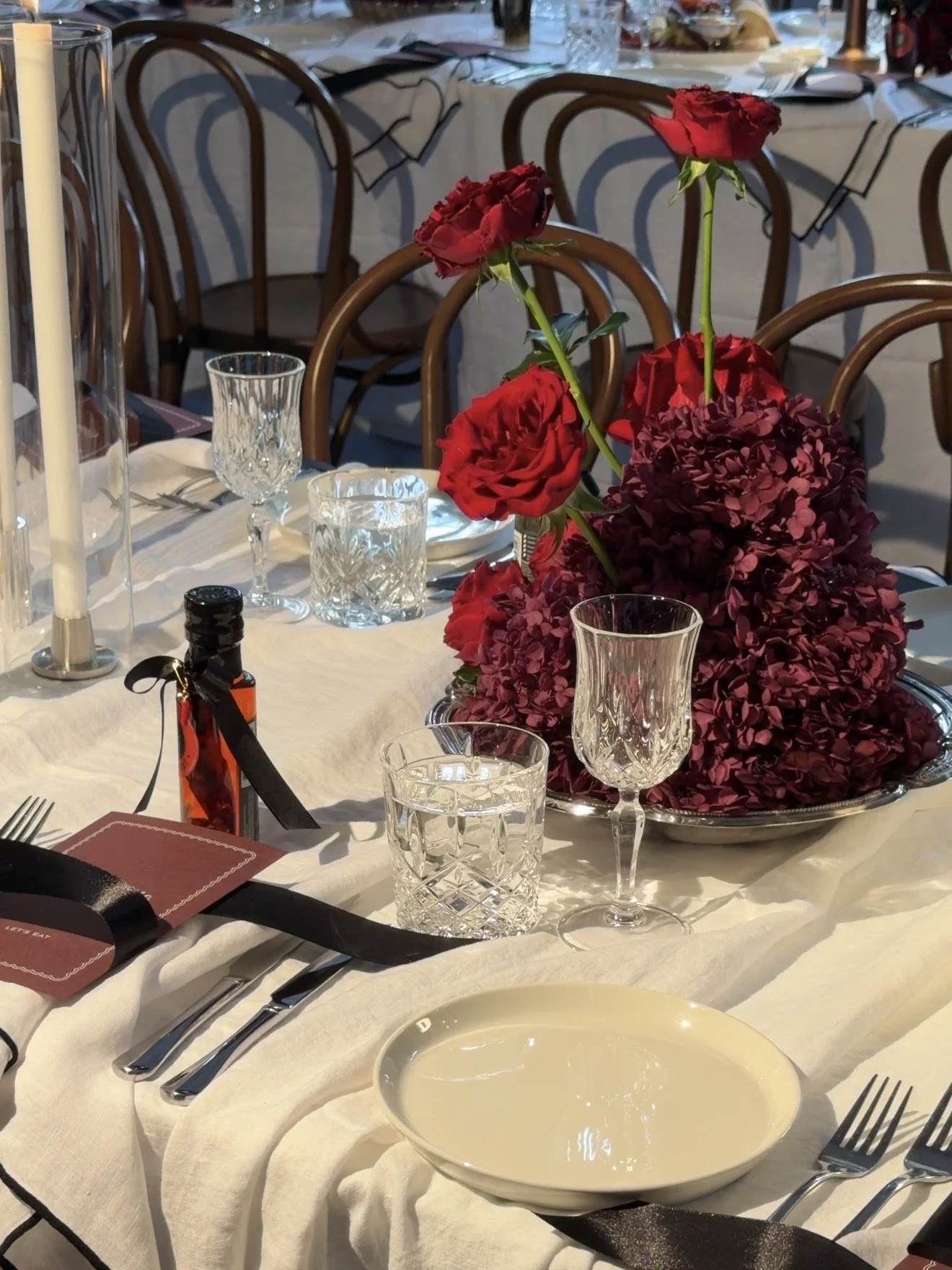 A decorated dining table with a floral centerpiece of red roses and purple hydrangeas, glassware, cutlery, and a small bottle of hot sauce, set for a formal meal.