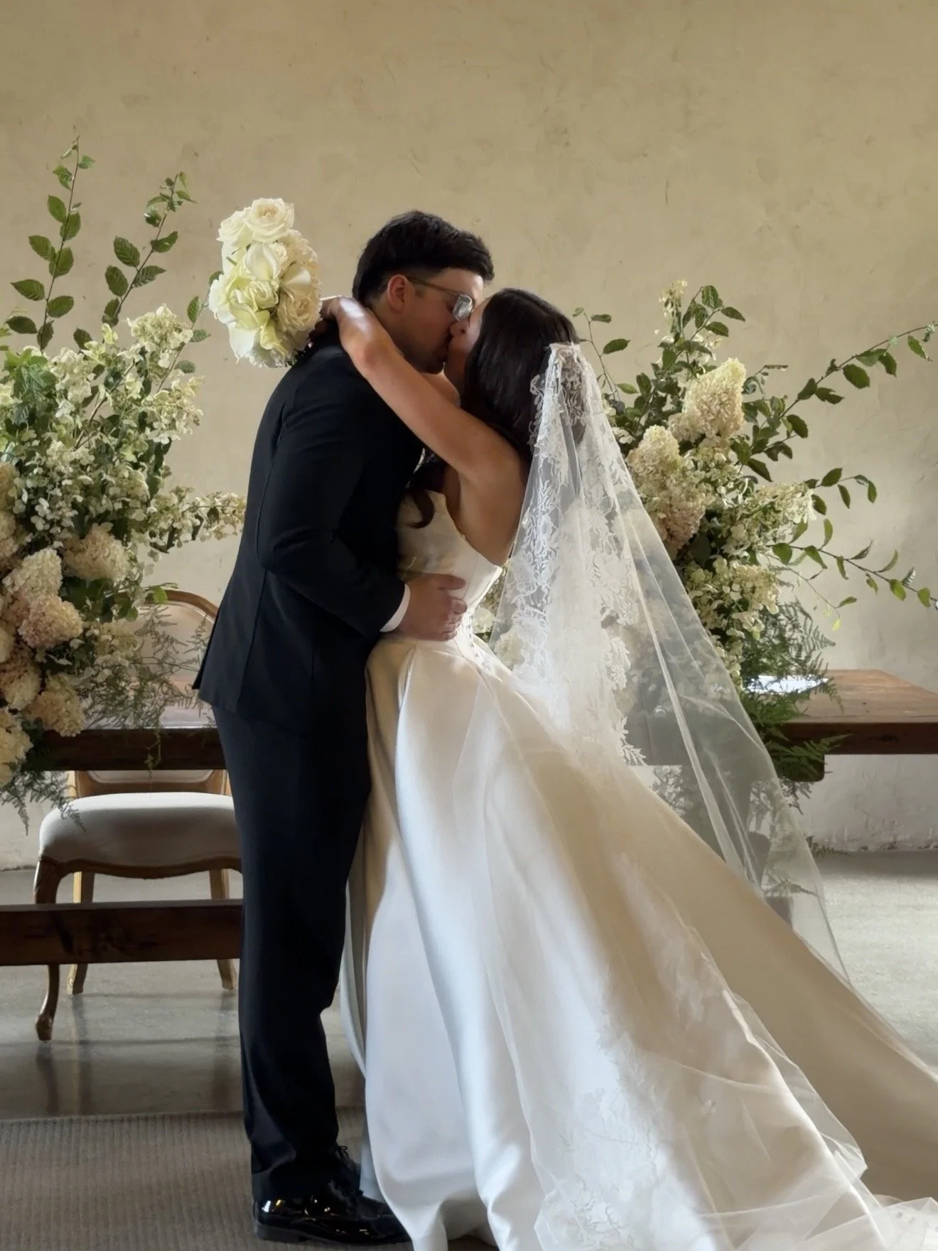 A bride and groom share a kiss during their wedding ceremony, surrounded by floral arrangements and seated chairs.