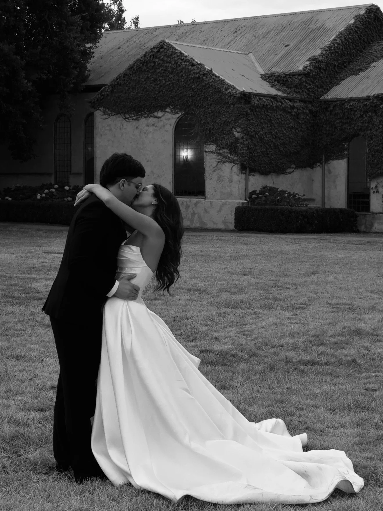 A bride and groom kiss outdoors on grass, the groom wearing a tuxedo and the bride in a strapless wedding gown.