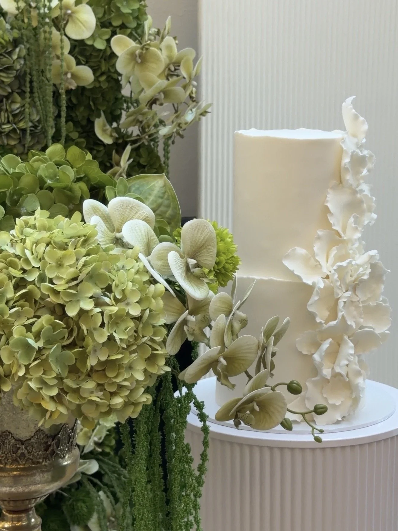 A white wedding cake decorated with white orchid flowers and creamy ruffled accents, surrounded by green floral arrangements at a celebration or wedding event.