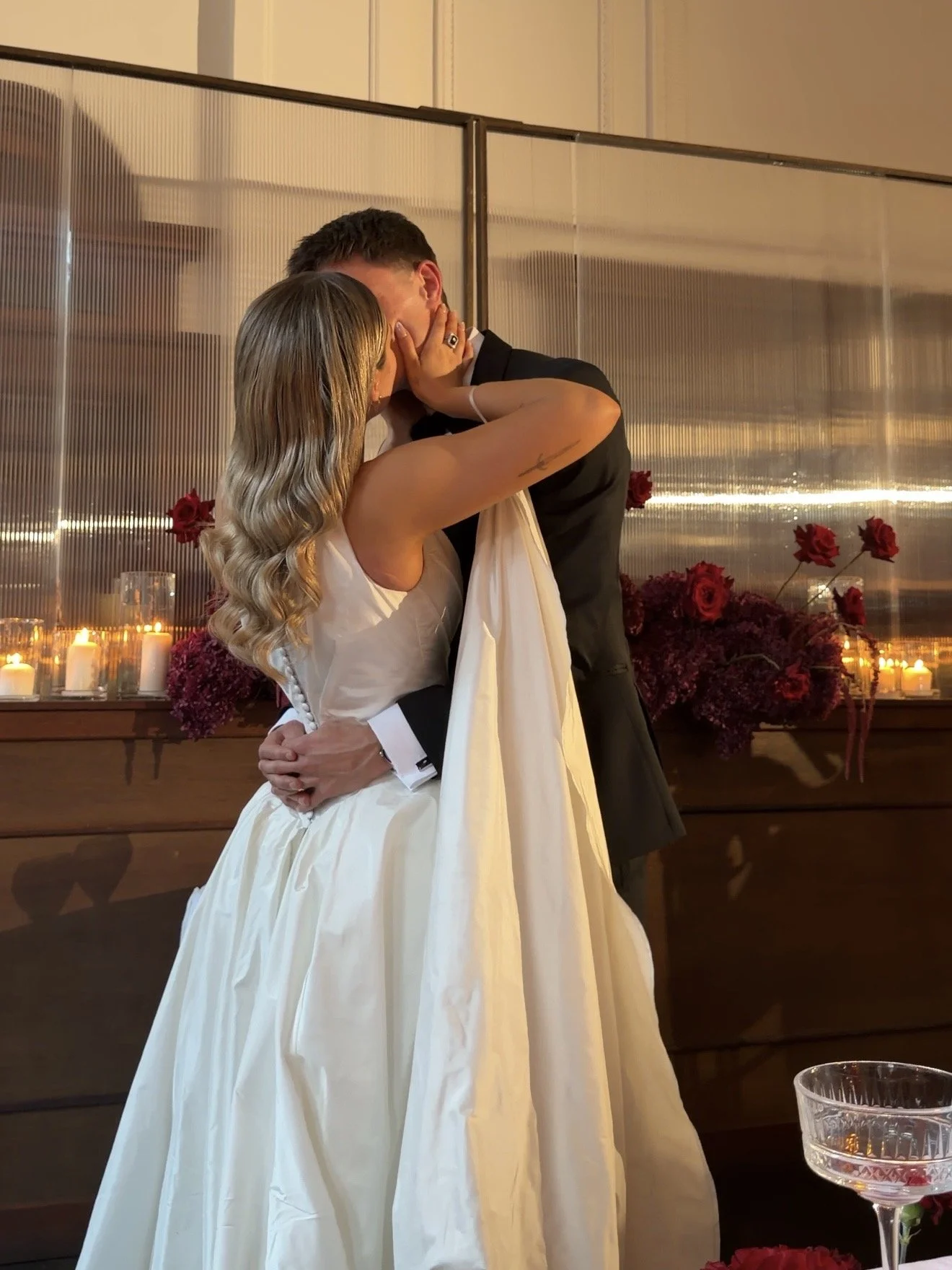 A bride and groom are kissing during their wedding, with the bride in a white gown and the groom in a black suit, inside a decorated venue with candles and red flowers.