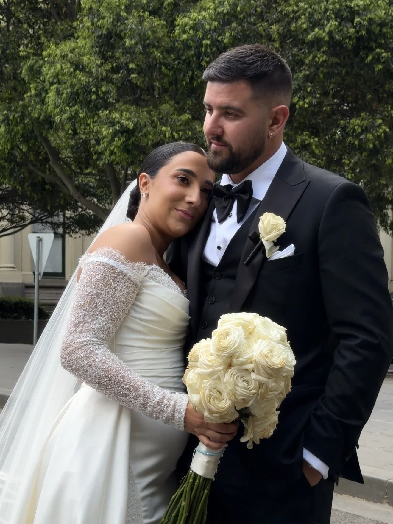 A bride and groom embracing, standing outdoors in front of green trees. The bride is holding a bouquet of white roses, and the groom is wearing a black tuxedo with a bow tie and a white rose boutonniere.