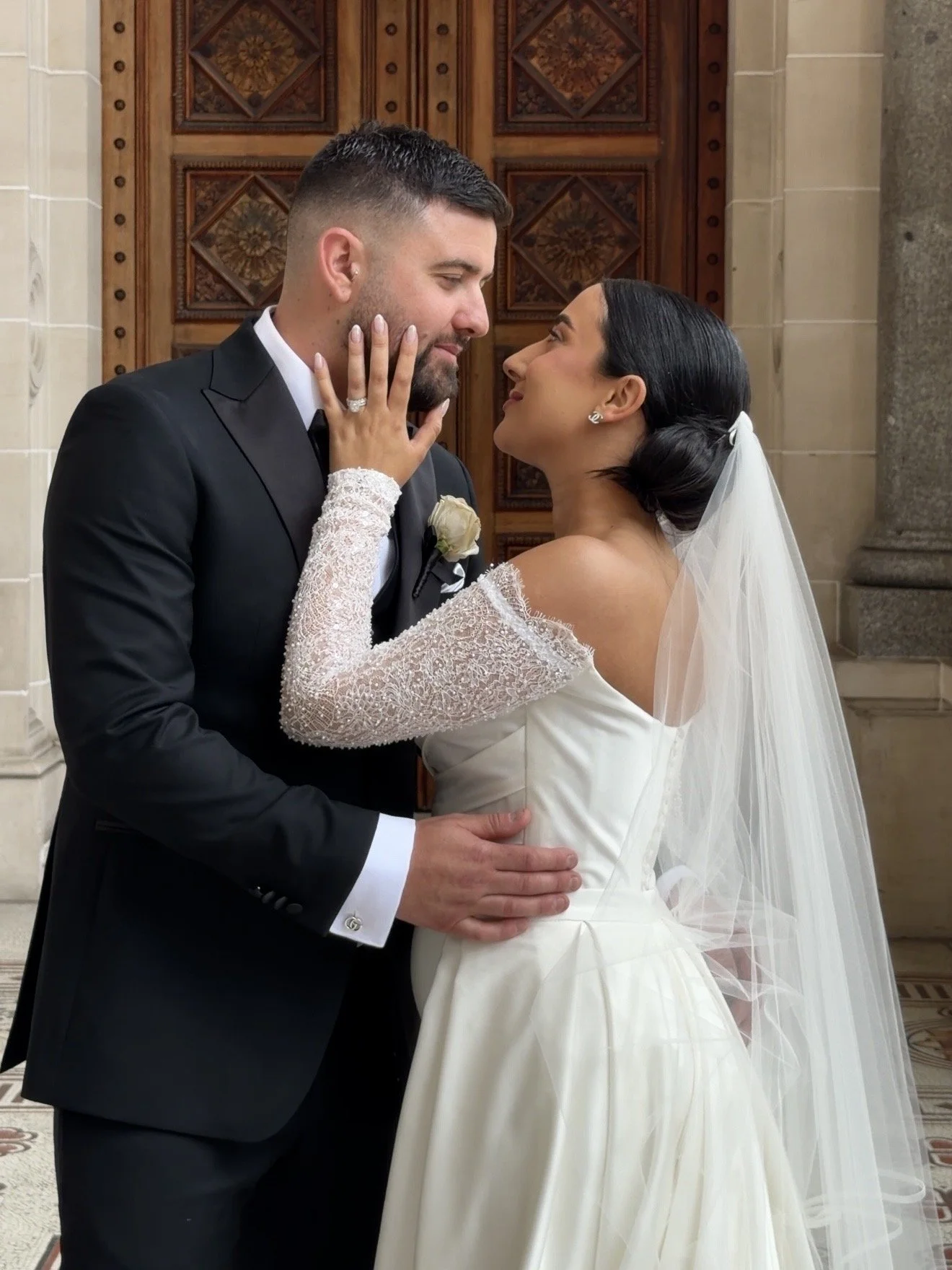 A bride and groom in a church, standing close and gazing at each other. The bride is wearing a white wedding gown with lace sleeves and a veil, and has dark hair styled in an updo. The groom is in a black tuxedo with a white shirt and boutonniere, ho