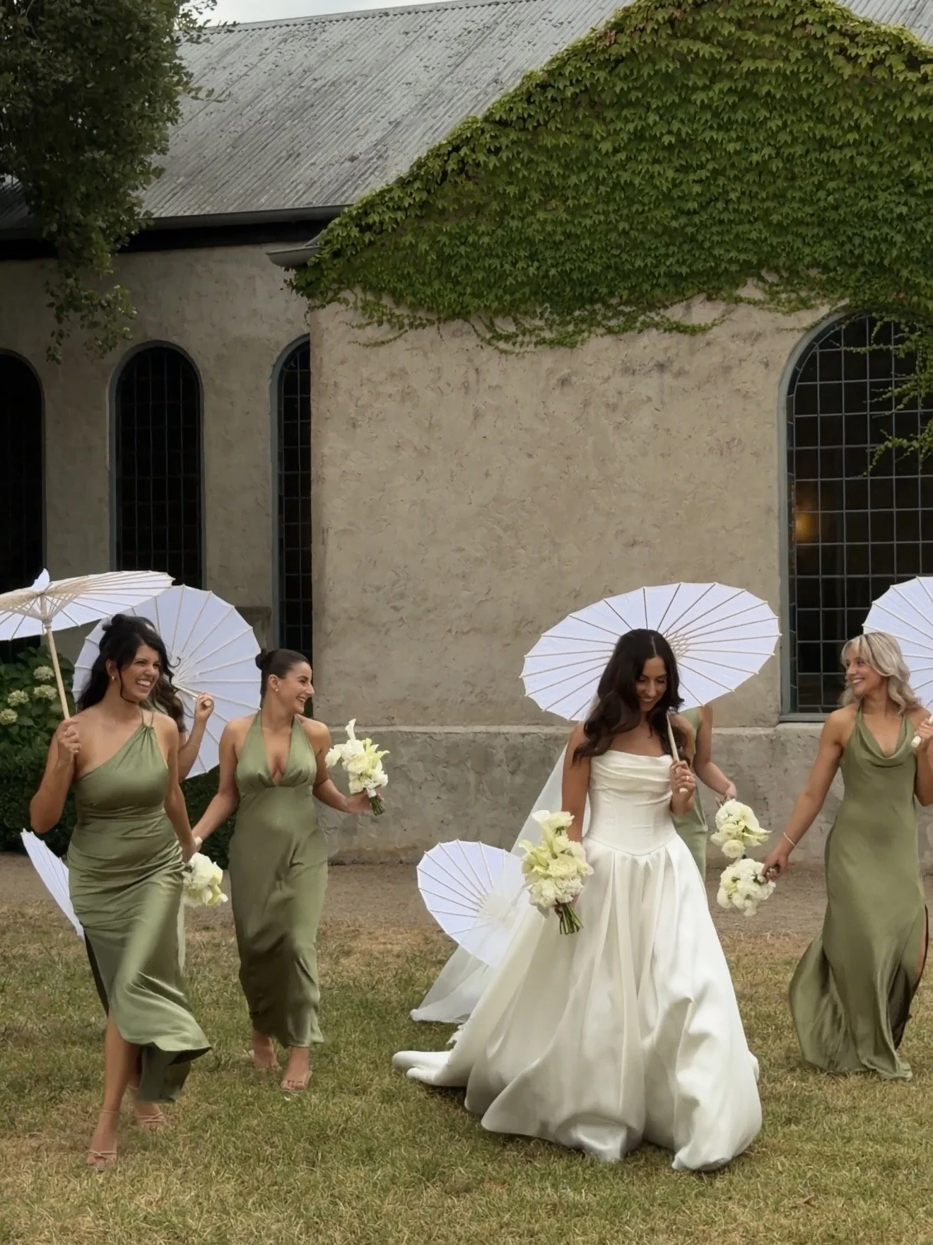 Bride in a white wedding dress walking on grass with four bridesmaids in matching olive green dresses, holding white umbrellas and bouquets, outdoors near a stone building covered in green ivy.