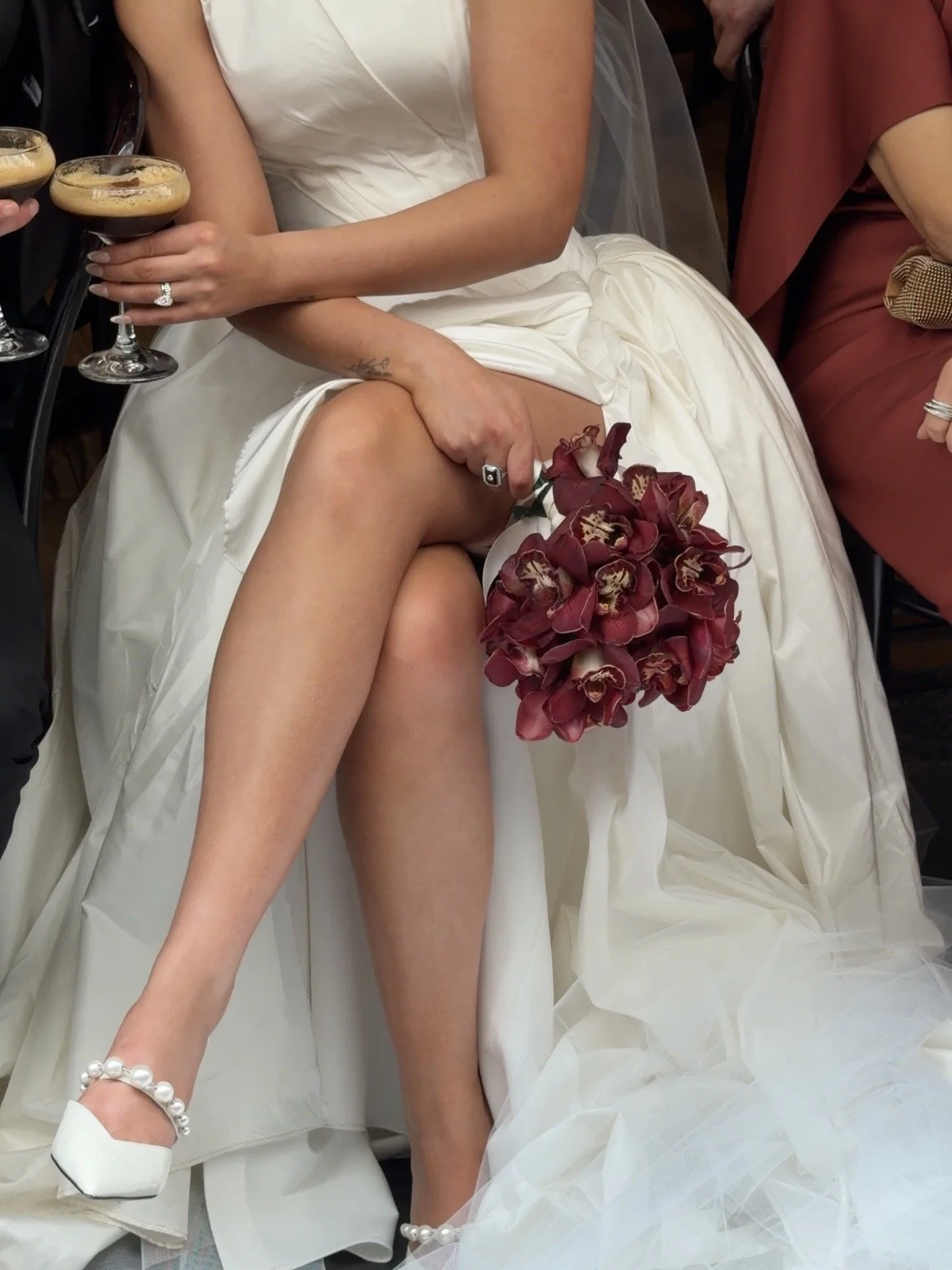 A bride in a white wedding dress is sitting, holding a bouquet of dark red flowers, with her left leg raised slightly, revealing a white wedding shoe with pearl embellishments. She is surrounded by guests holding drinks at a celebration.