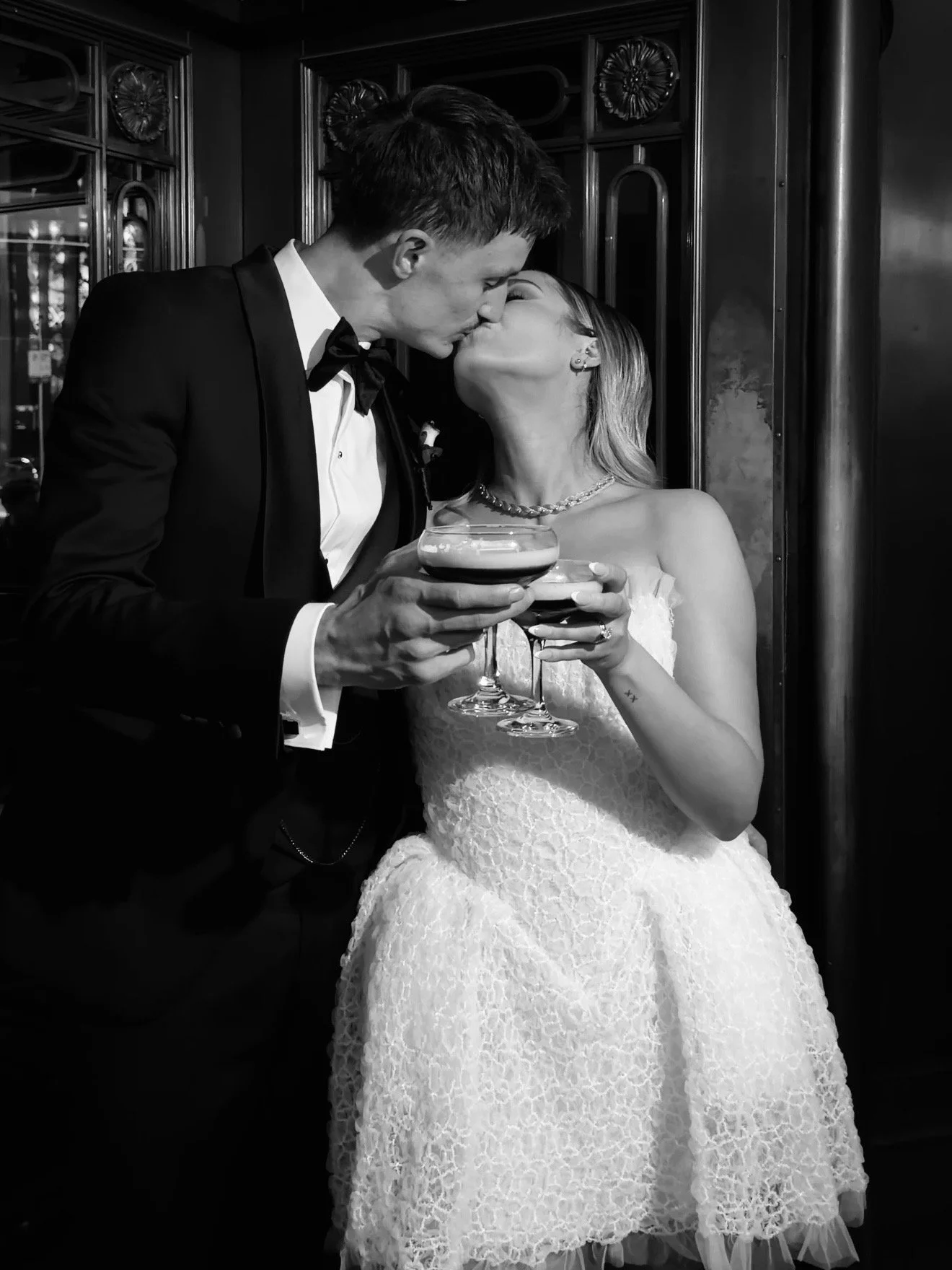 A couple in wedding attire sharing a kiss while holding two espresso martinis in a vintage-themed setting.