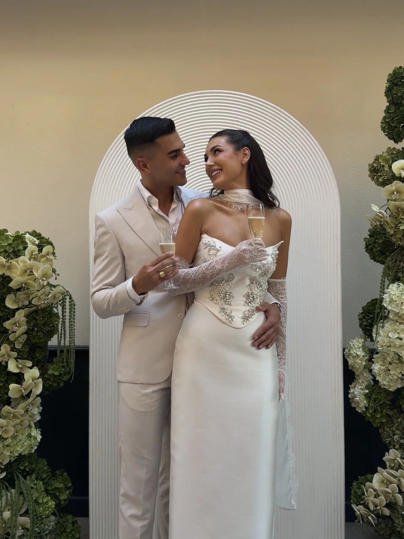 A bride and groom holding champagne glasses, smiling at each other, standing in front of an arch with floral decorations.