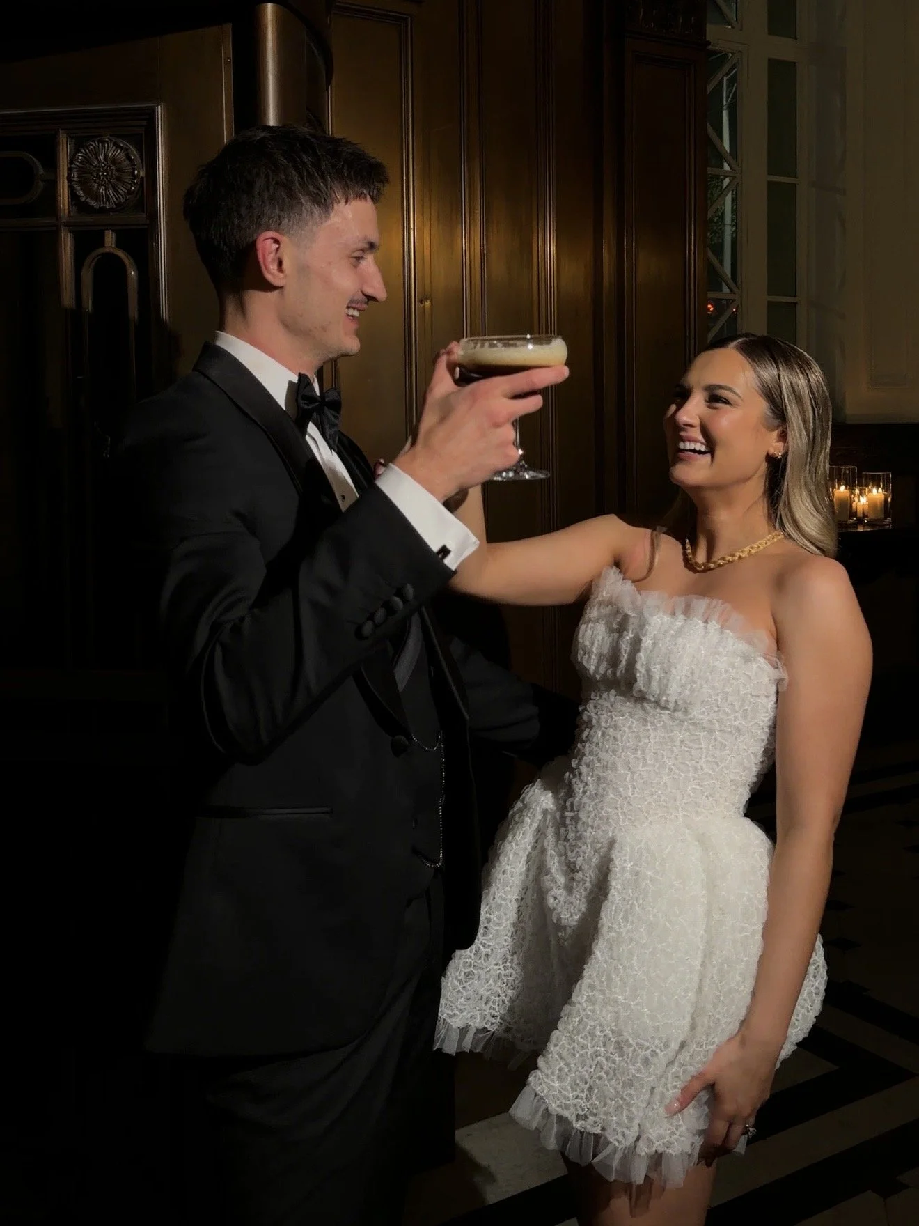 A man in a tuxedo and a woman in a white dress smiling at each other as the man holds up a cocktail glass.