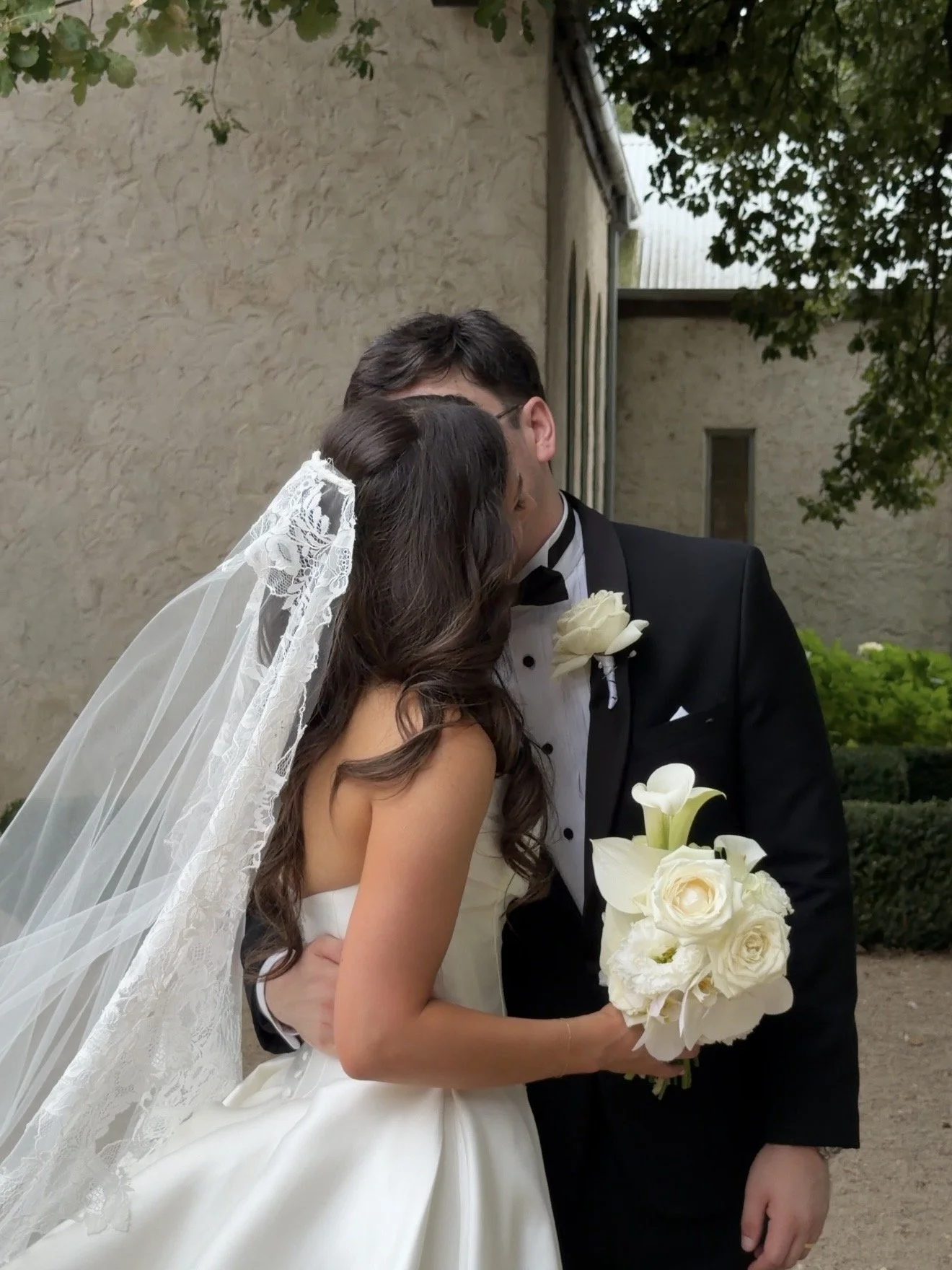 A bride and groom share a kiss outdoors, the bride in a white wedding dress with a lace veil and holding a bouquet of white flowers, the groom in a black tuxedo with a white boutonniere, against a rustic wall background.