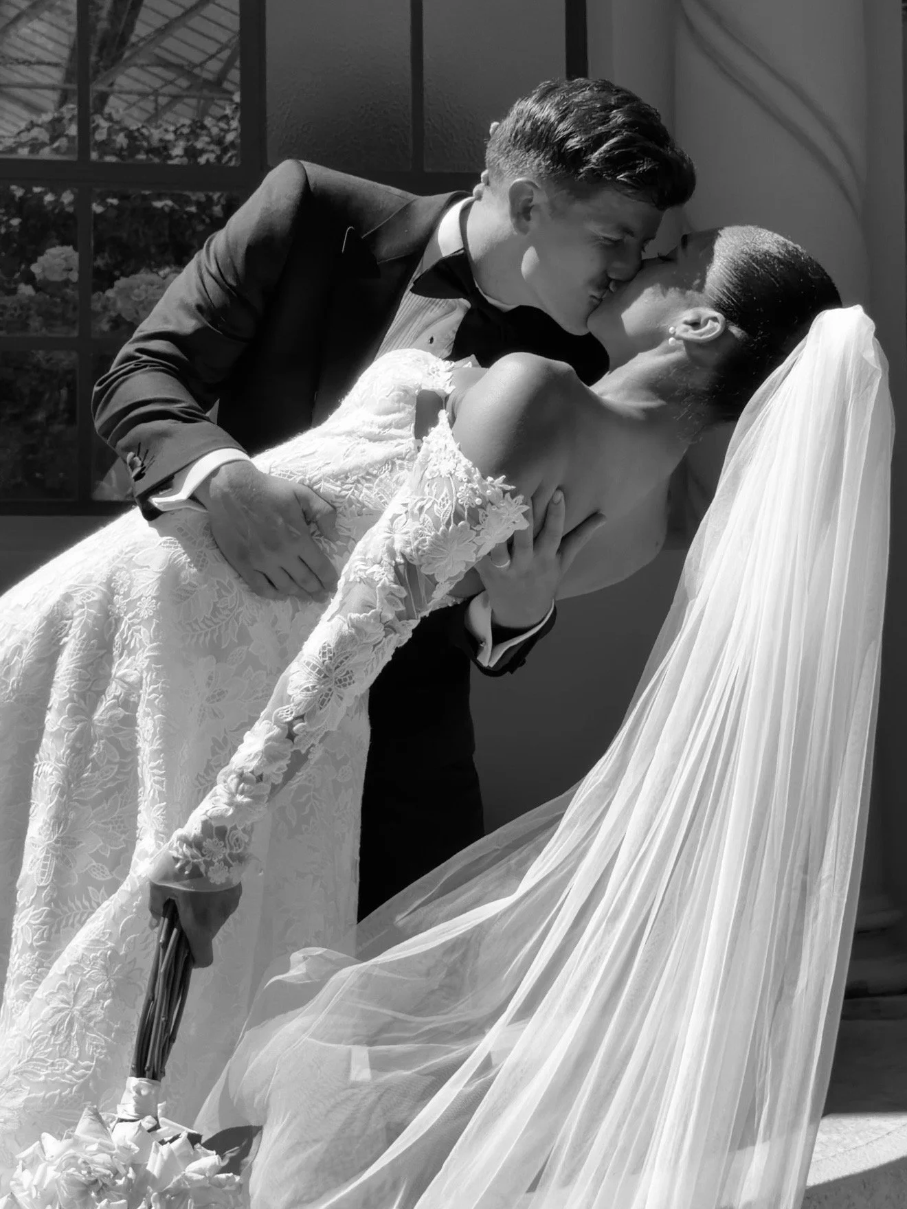 A couple in wedding attire sharing a kiss, with the groom in a tuxedo and the bride in a lace wedding dress and veil, during a wedding ceremony.